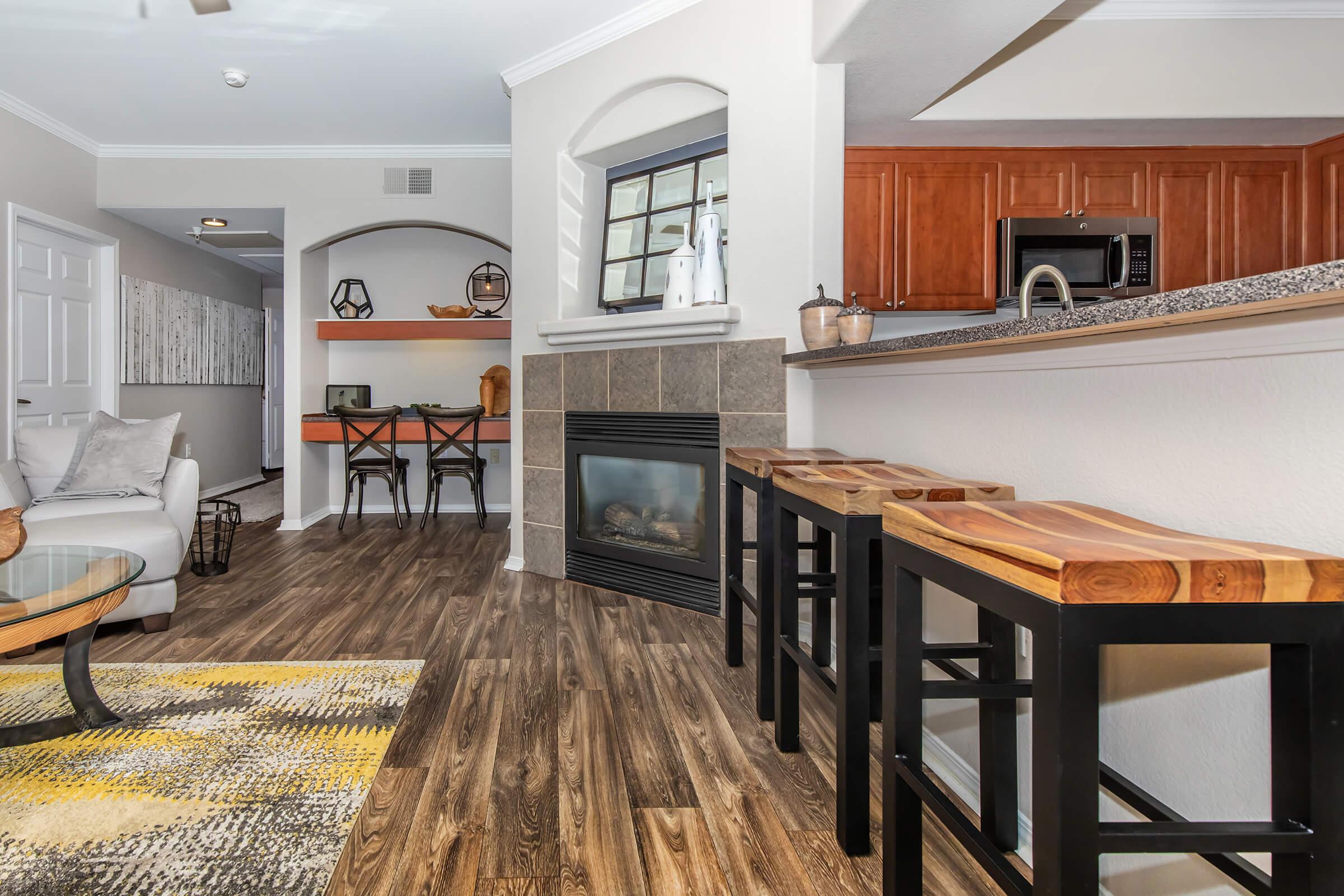 A modern living space featuring a cozy seating area with a white couch, wooden bar stools at a kitchen countertop, and a fireplace. Light wood flooring complements the warm tones of the cabinetry and decor, with a dining table visible in the background, creating an inviting atmosphere.