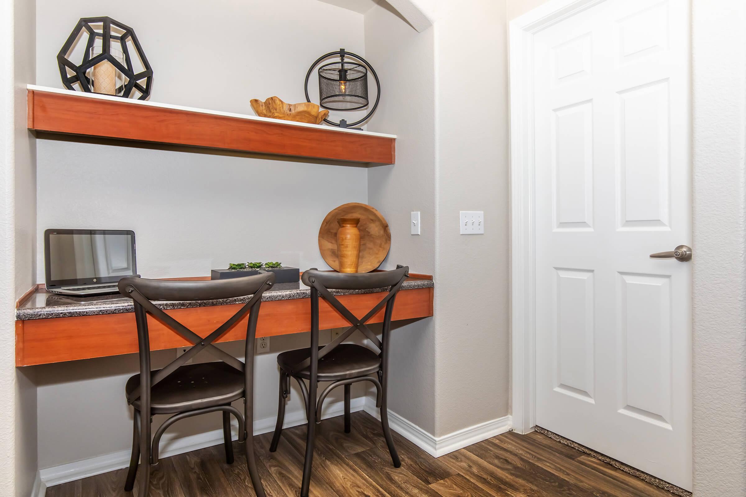 A small home office nook featuring a wooden desk with two black chairs, a laptop on the desk, decorative items above, and a door in the background. The walls are painted light gray, and the floor has a wood-like finish, creating a cozy and modern workspace.