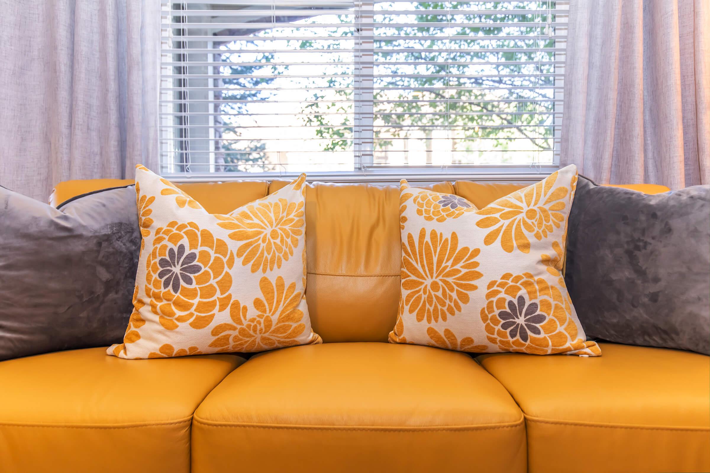A bright yellow sofa with two decorative pillows. One pillow has a floral pattern in shades of yellow and gray, while the other is a solid gray. The backdrop includes curtains and a window with greenery outside, creating a cheerful and inviting living space.