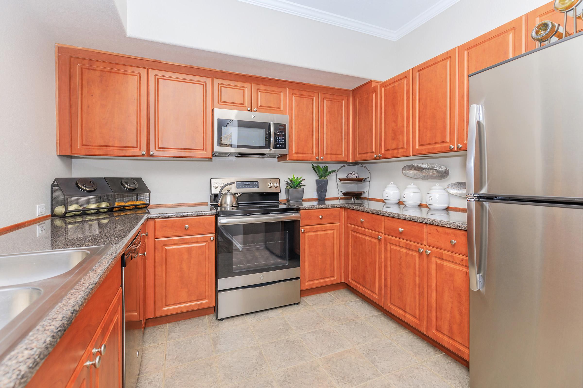 A modern kitchen featuring wooden cabinets, a stainless steel refrigerator, an oven, and a microwave. The countertop is made of granite, and there are decorative items including a plant and white dishware displayed on the shelves. The kitchen is well-lit and organized.