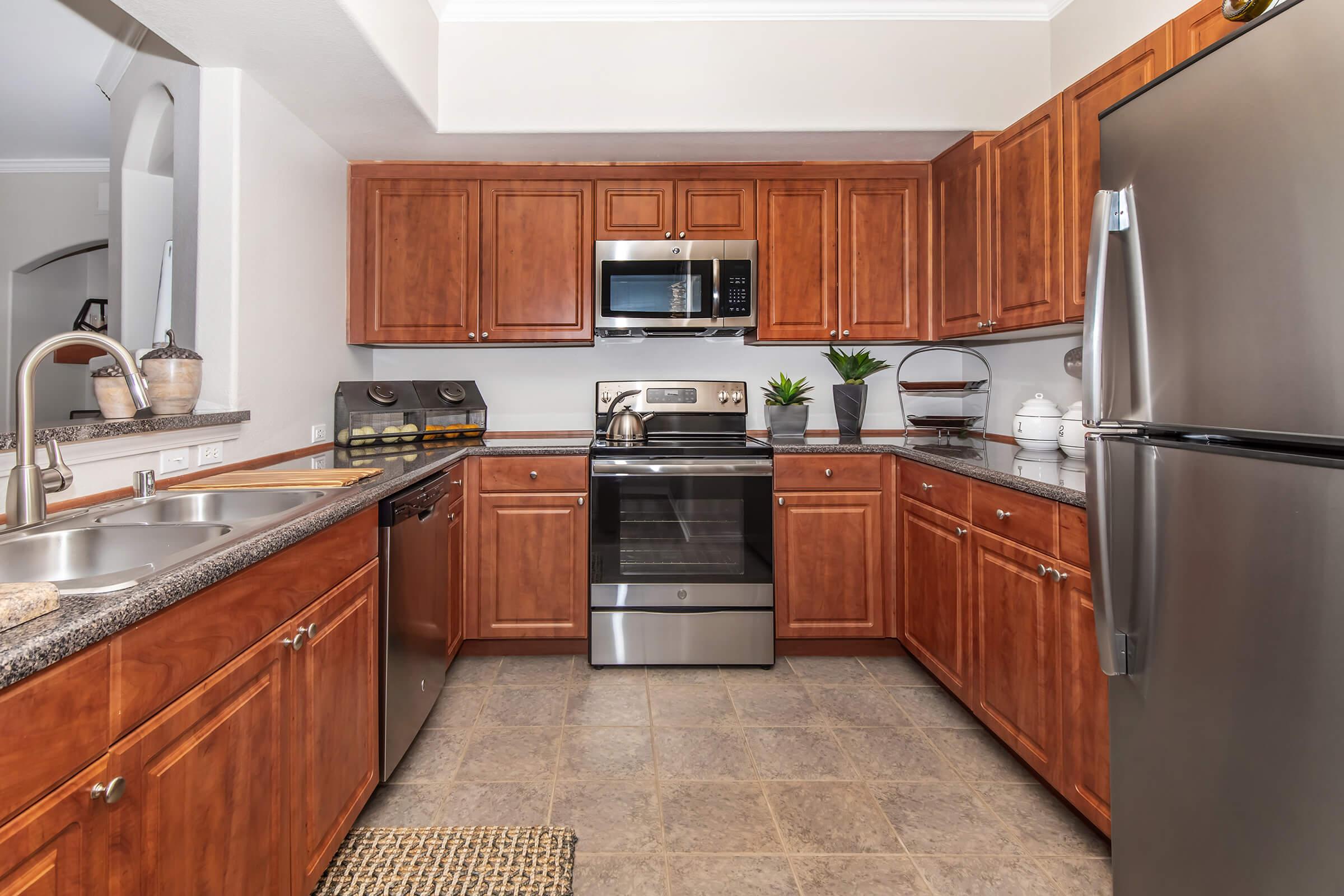A modern kitchen featuring wooden cabinetry, a stainless steel refrigerator, and an oven with a microwave above. The countertop is granite, and there are decorative items like plants and a dish rack. The floor is tiled, and a woven rug is placed near the sink.