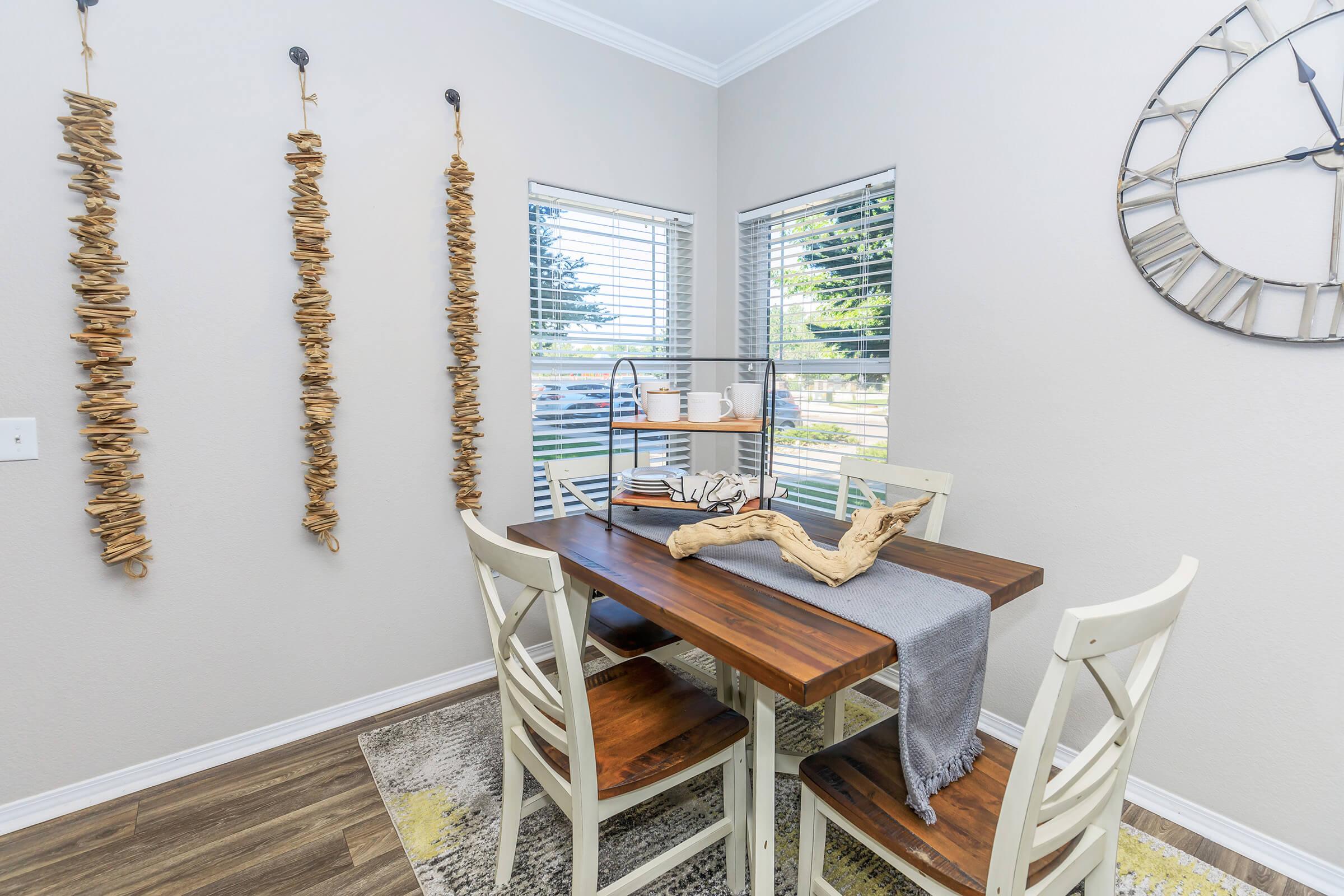 A cozy dining area featuring a wooden table with a gray table runner and rustic decor. There are two white chairs, a decorative wooden piece on the table, and a stylish shelf with dishes. Three vertical decorations made of natural wood hang on the wall, and a large wall clock is visible. Sunlight streams through the windows.