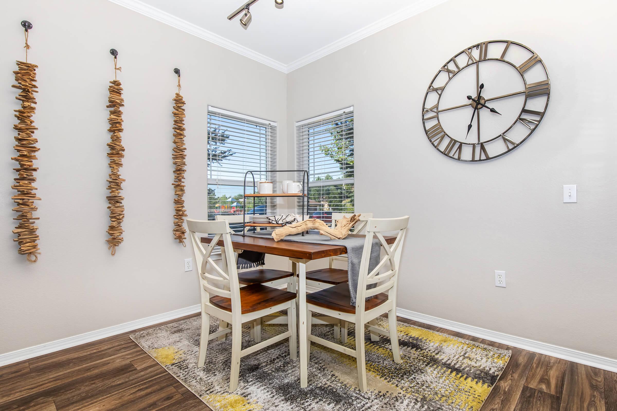A cozy dining area featuring a wooden table with four chairs, adorned with a decorative centerpiece. The walls are decorated with hanging wooden art pieces, and a large round clock is mounted above. Natural light streams through two windows, enhancing the inviting atmosphere. A patterned rug adds warmth to the space.