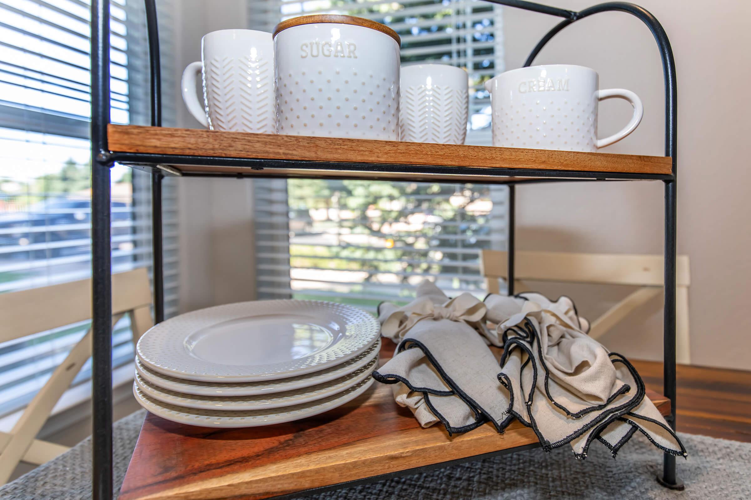 Two-tier wooden serving cart with white ceramic cups labeled "SUGAR" and "CREAM," and a stack of white plates below. A set of neatly folded napkins with contrasting edges sits beside the plates. Natural light filters through window blinds, creating a cozy, inviting atmosphere.