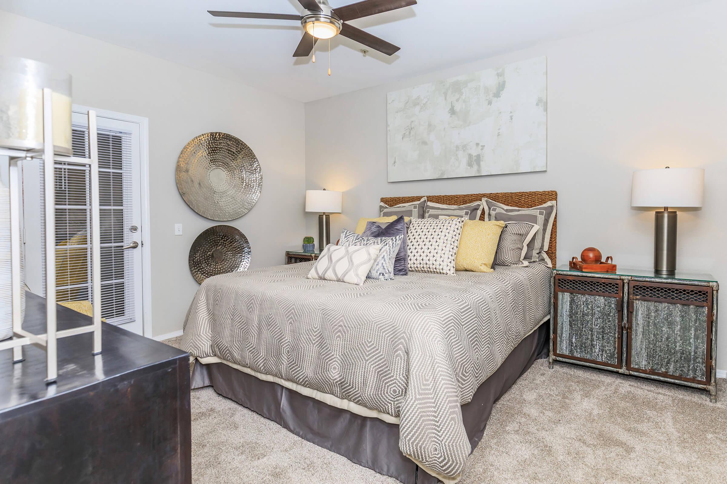 A stylish bedroom featuring a king-sized bed with decorative pillows, a light gray comforter, and a large piece of abstract art above the headboard. Two lamps on nightstands provide soft lighting, and there are metallic wall decor pieces. A dark wooden dresser and soft carpet complete the cozy atmosphere.