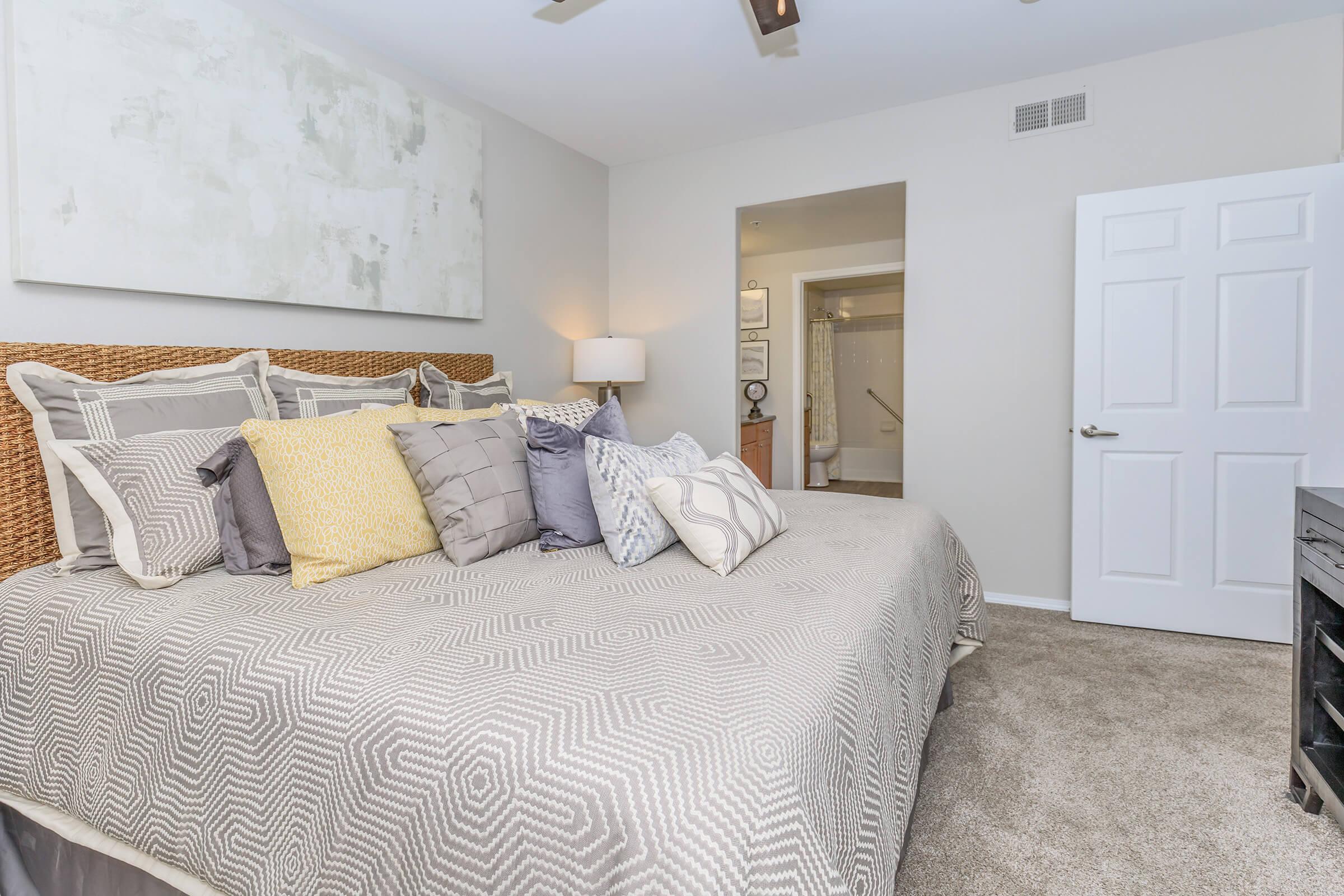 A stylish bedroom featuring a large bed with decorative pillows, a textured gray blanket, and a neutral color palette. The walls are painted in soft tones, with a large piece of abstract wall art above the bed. A lamp beside the bed adds warm lighting, and a door in the background leads to a bathroom.