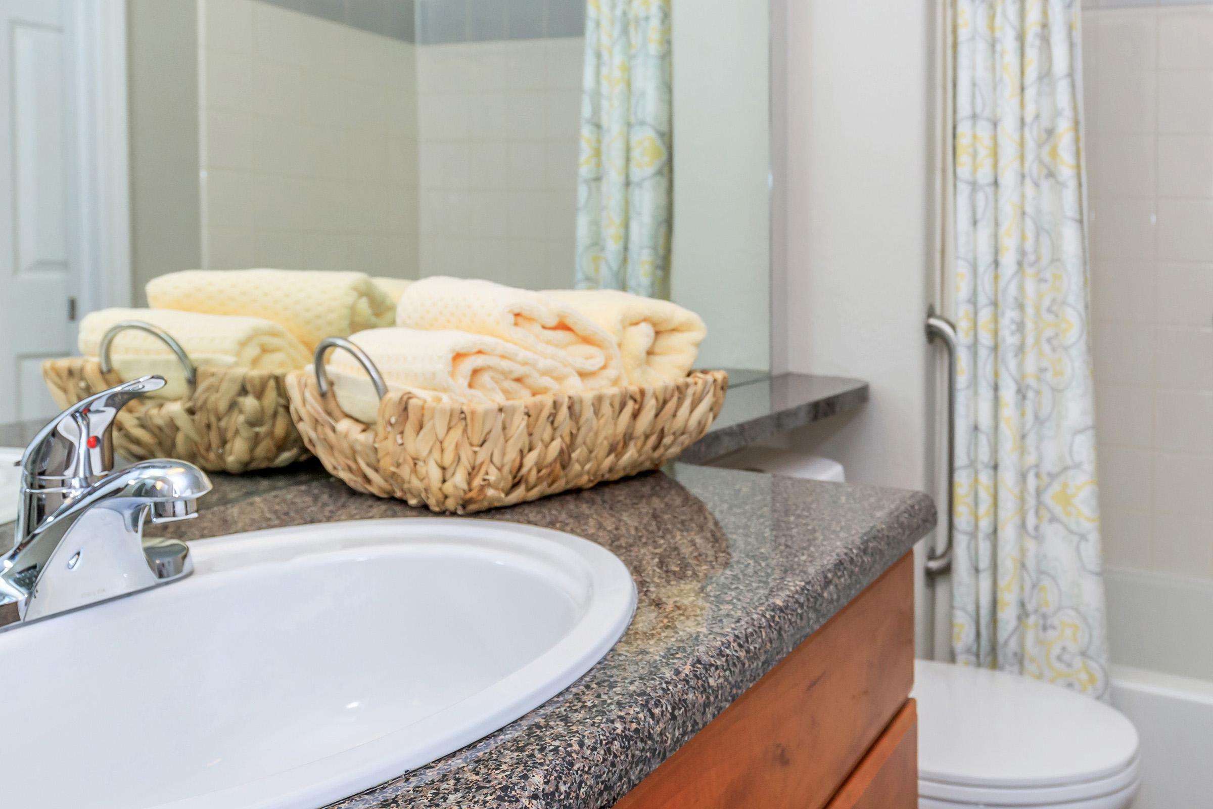 A clean bathroom countertop featuring a sink, a faucet, and a woven basket filled with neatly rolled yellow towels. In the background, there is a shower curtain with a subtle pattern and a toilet partially visible. The space is well-lit and organized, contributing to an inviting atmosphere.