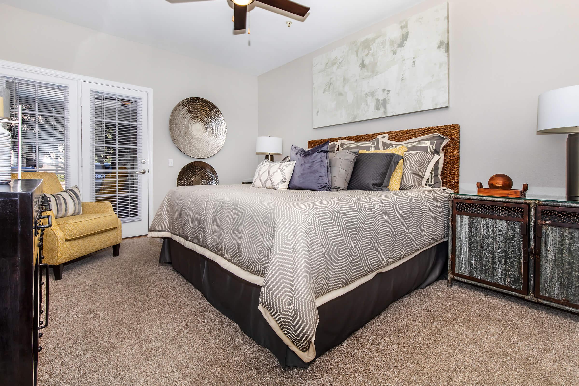 A cozy bedroom featuring a large bed with decorative pillows, a neutral-colored bedspread, and stylish wall art. There's a comfortable chair in the corner, modern bedside lamps, and a textured cabinet. Light streams through the windows, creating a warm and inviting atmosphere.
