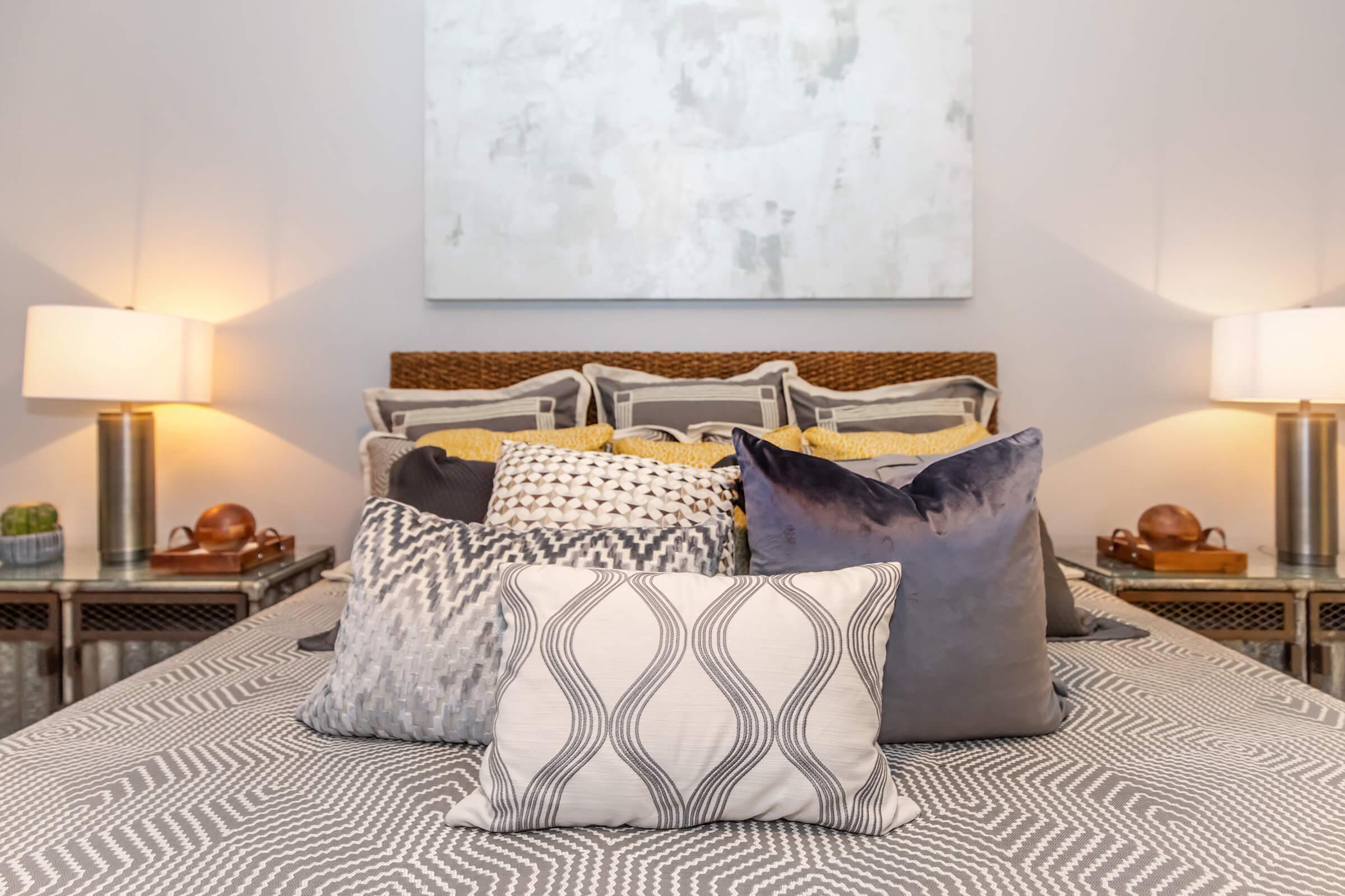 A beautifully decorated bedroom featuring a bed adorned with various decorative pillows in shades of gray, yellow, and purple. Soft table lamps are placed on either side of the bed, providing warm lighting, while a large abstract artwork hangs above the headboard. The bedding has a subtle geometric pattern.