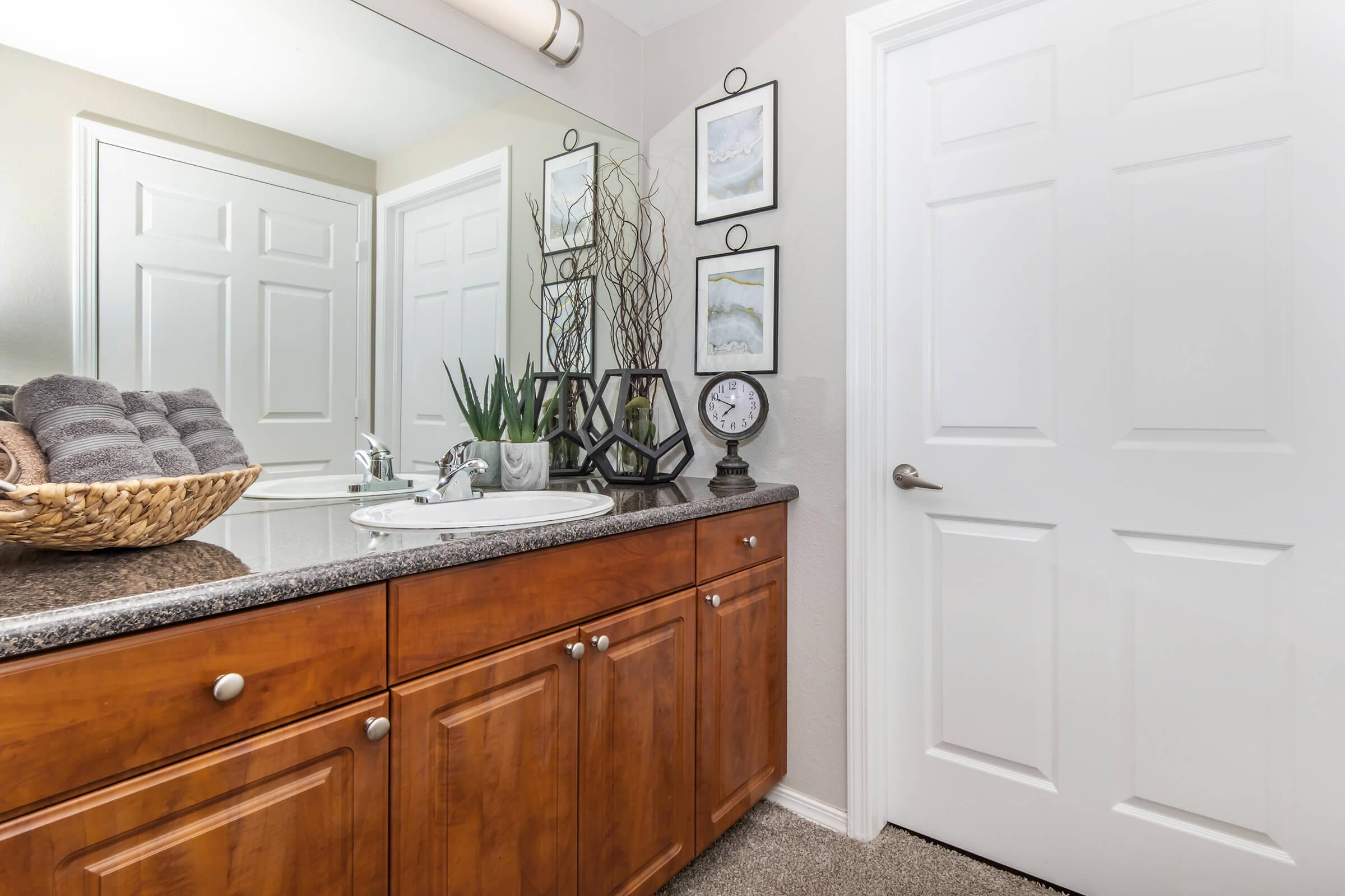 A modern bathroom featuring a granite countertop with a sink, stylish decor including potted plants and framed art on the wall, a clock on the countertop, and neatly stacked towels in a woven basket. The design is complemented by a mirror reflecting the space, highlighted by a light color palette.