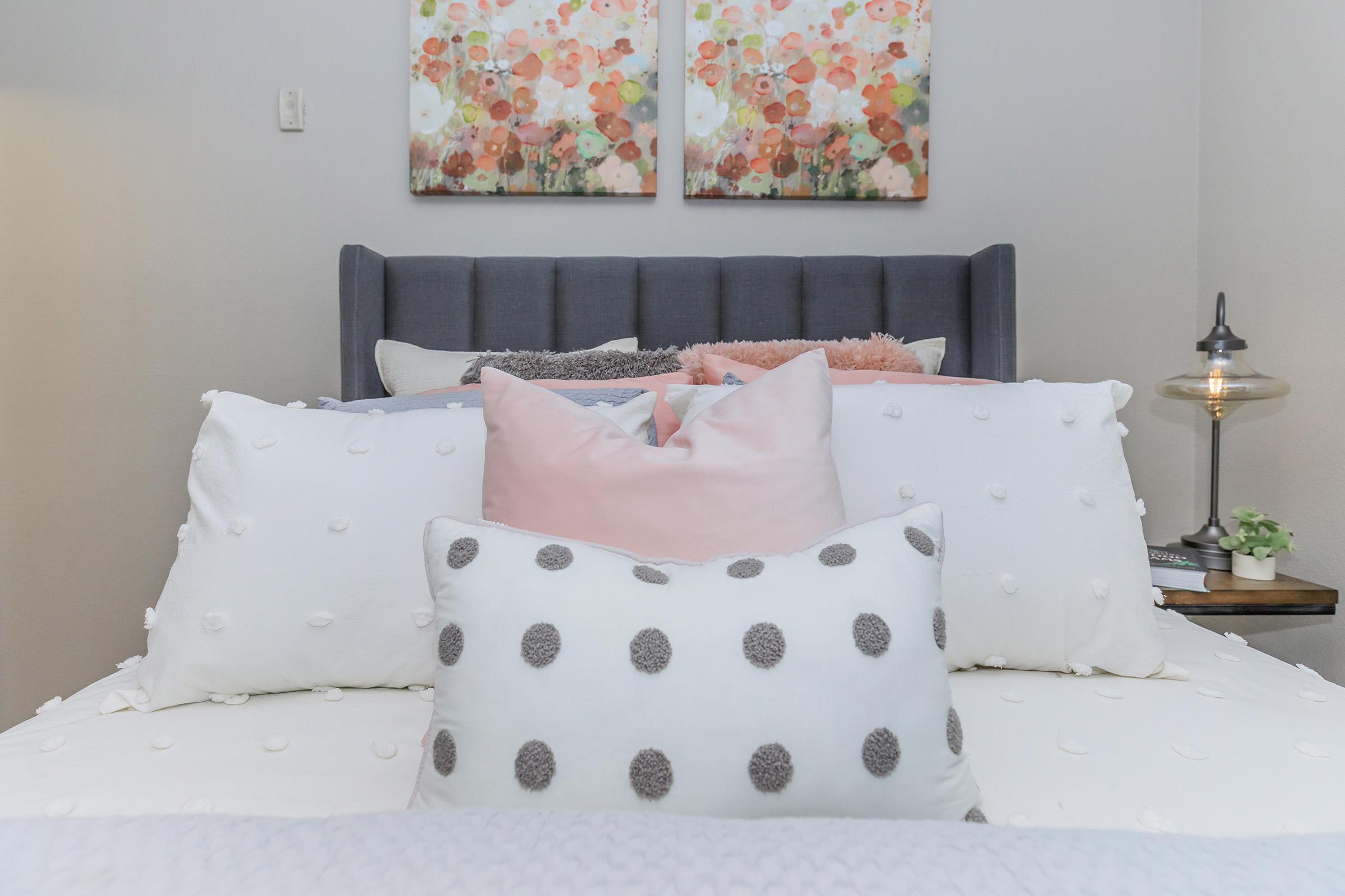 A cozy bedroom scene featuring a neatly made bed with multiple decorative pillows, including a white pillow with polka dots and a pink one. The headboard is gray, and two floral paintings hang on the wall above. A bedside table with a lamp and a small green plant adds warmth to the setting.