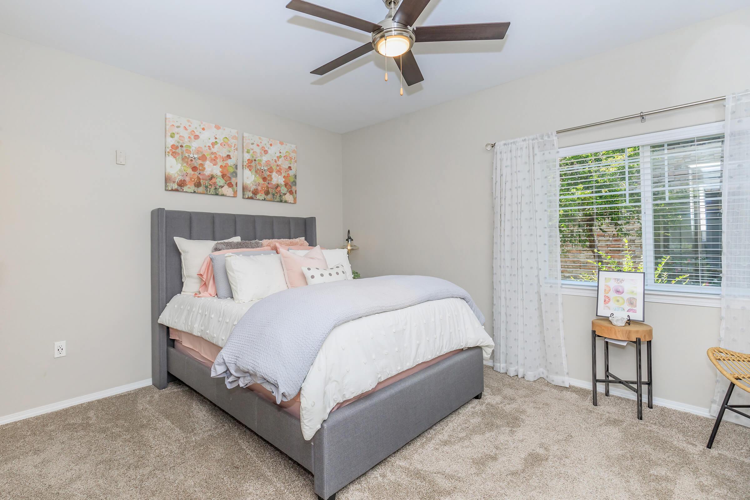 A cozy bedroom featuring a gray upholstered bed with decorative pillows and a soft blanket. Natural light enters through a window adorned with sheer curtains, and a floral painting hangs above the bed. A bedside lamp and a small wooden table with decor complete the inviting atmosphere. Soft beige walls and carpet enhance the warmth.
