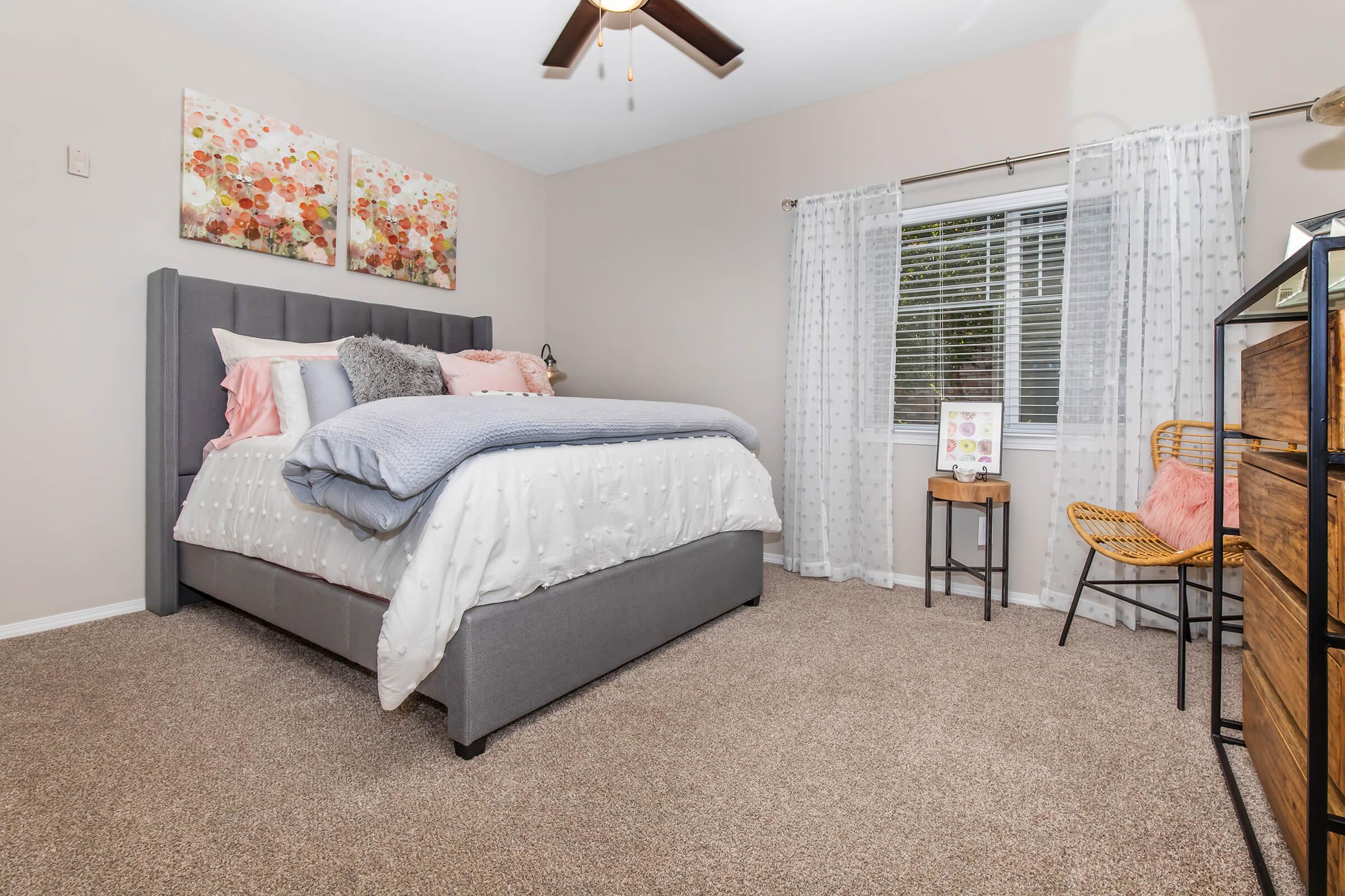 A cozy bedroom featuring a gray upholstered bed with a light pink and gray comforter. Two floral wall paintings hang above the bed. A window with sheer curtains lets in natural light, and there's a small wooden nightstand with a decorative piece next to a rattan chair. The carpeted floor adds warmth to the space.