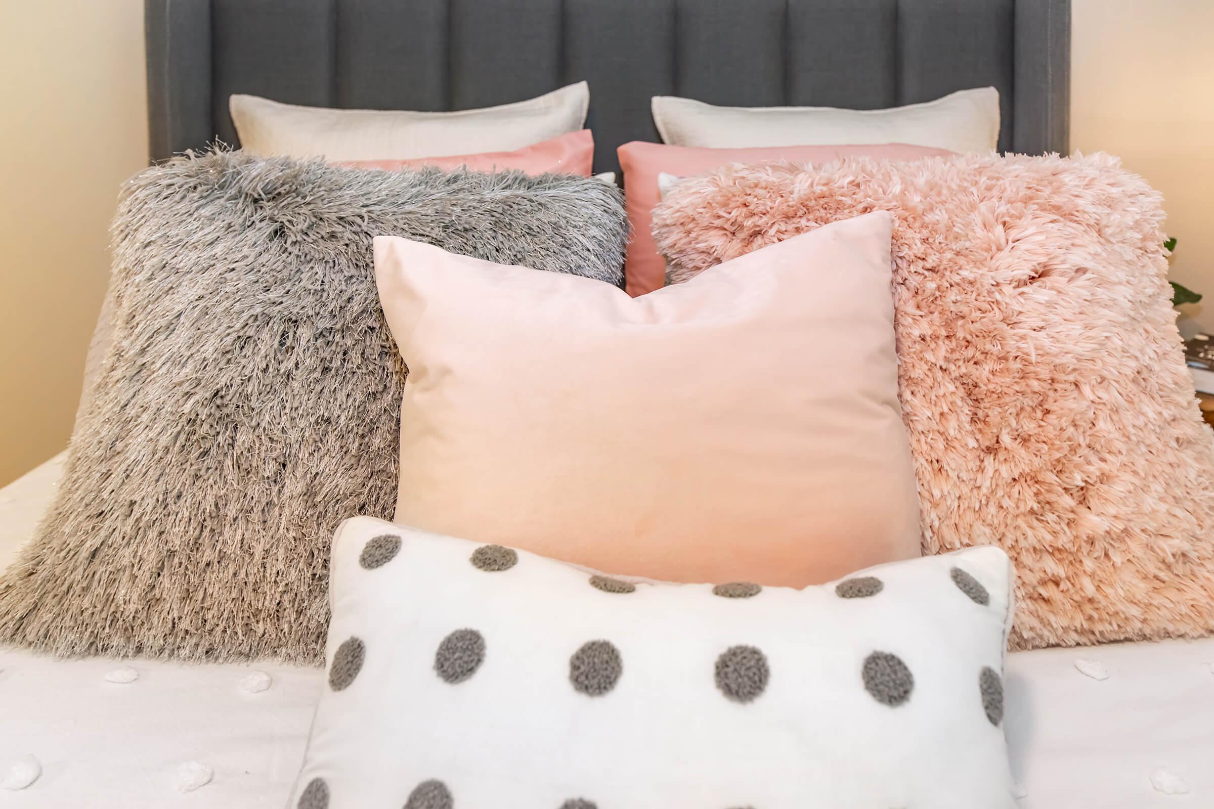 A cozy arrangement of decorative pillows on a bed. The pillows vary in texture and color, featuring soft fur-like materials in gray, pink, and peach shades, along with a white pillow with gray polka dots. The backdrop includes a stylish gray headboard and softly lit bedside decor.