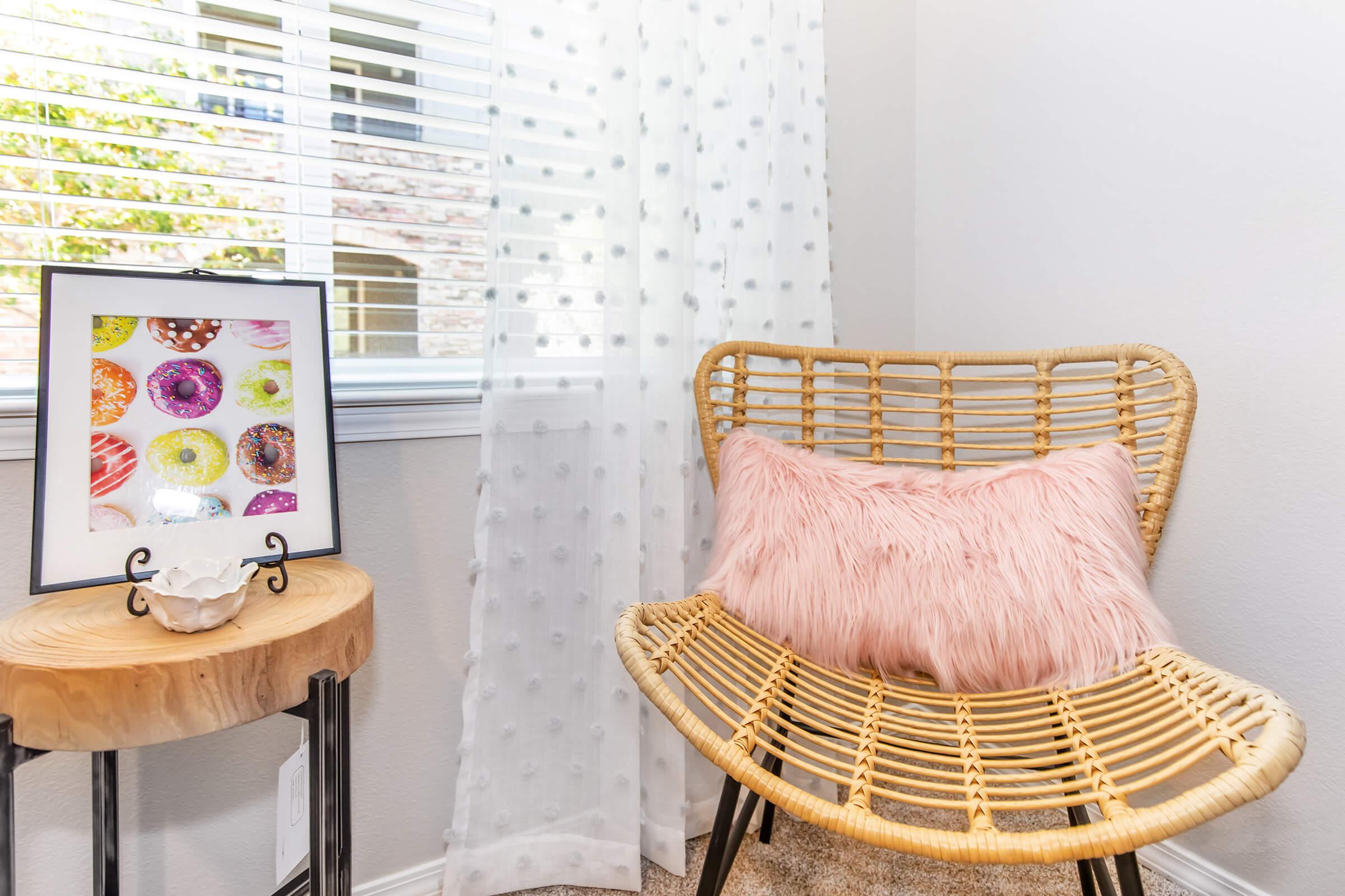 A cozy corner featuring a rattan chair with a fluffy pink pillow, next to a wooden side table holding a decorative shell. A framed colorful print of donuts is displayed on the table. Sheer white curtains softly filter natural light from a nearby window.