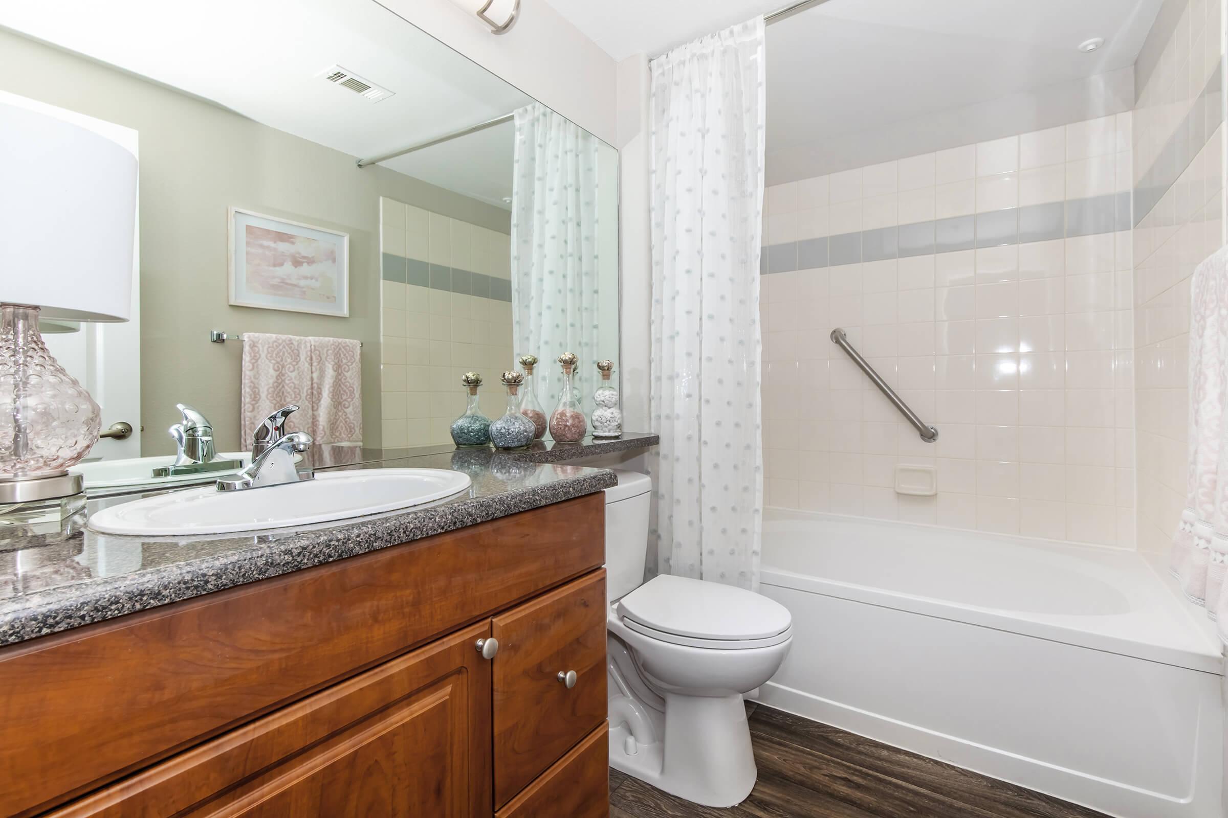 A modern bathroom featuring a white bathtub with a shower, a sink with a granite countertop, and light wood cabinetry. The space is decorated with a mirror, decorative bottles, and soft lighting from a lamp. Curtains provide privacy, and the overall color scheme is neutral and inviting.