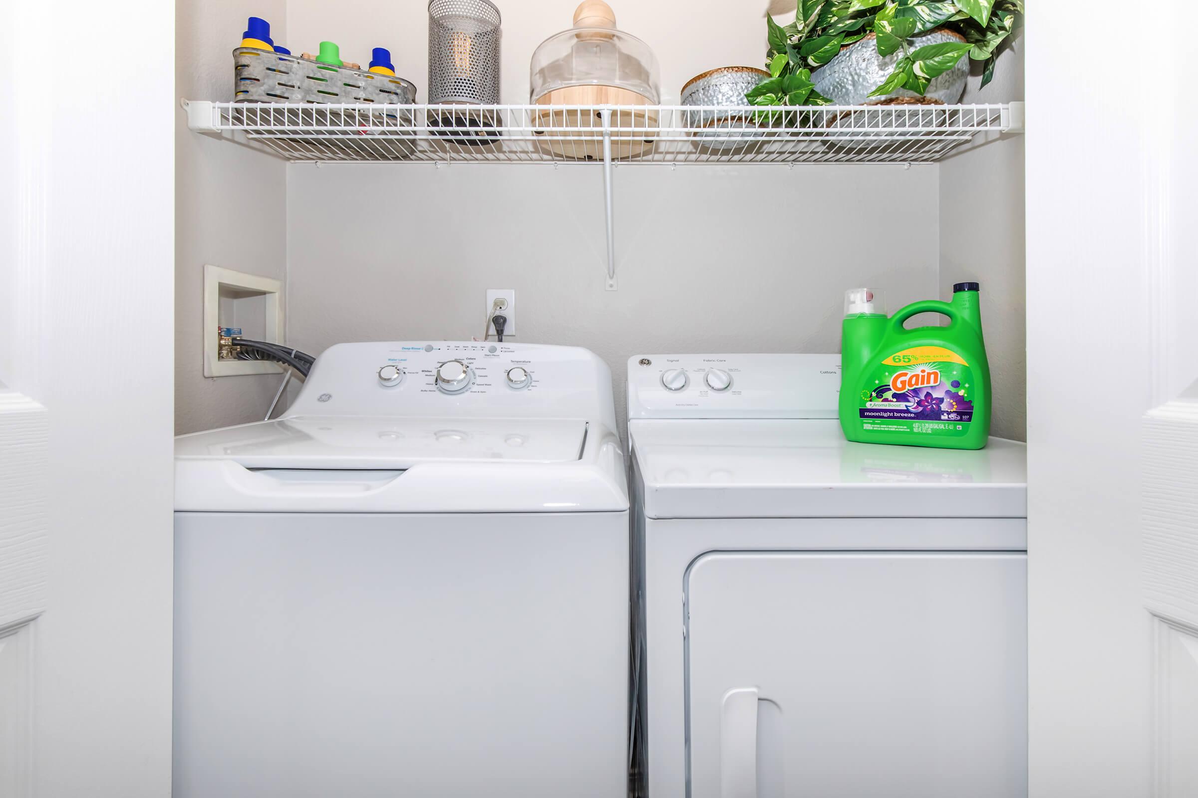 A laundry room featuring a white washing machine and dryer side by side on a shelf. Above them, there are various containers and a decorative plant. The wall is painted light gray, and the area is well-organized and clean. A bottle of Gain laundry detergent is prominently displayed on the right.