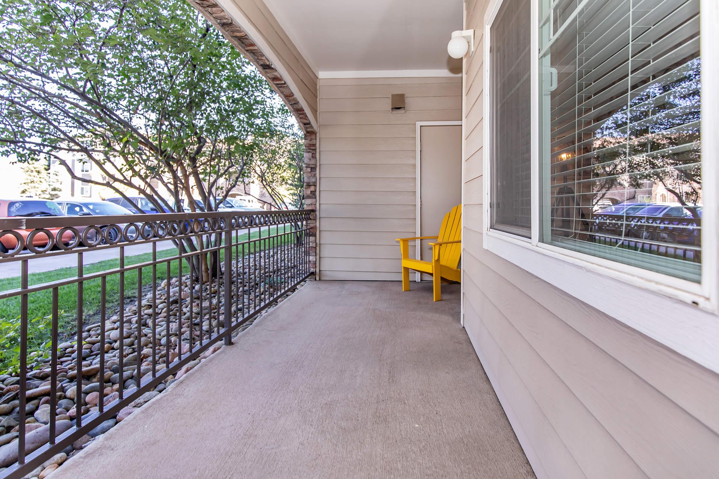 A front porch with a concrete floor and a yellow adirondack chair positioned beside a window. Decorative stone landscaping is visible in the foreground, and a path leads to a grassy area outside with a view of parked cars across the street.