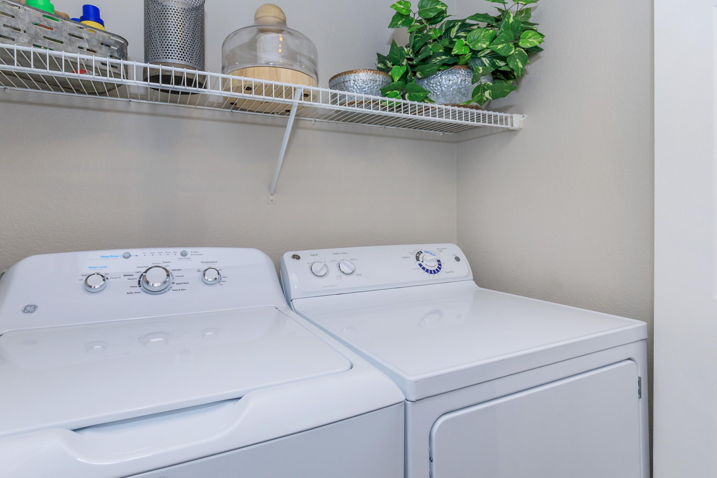 A laundry room featuring a white washing machine and a white dryer side by side. Above them is a wire shelf holding decorative items, including a plant and various containers. The wall is painted a light color, creating a clean and organized appearance in the space.