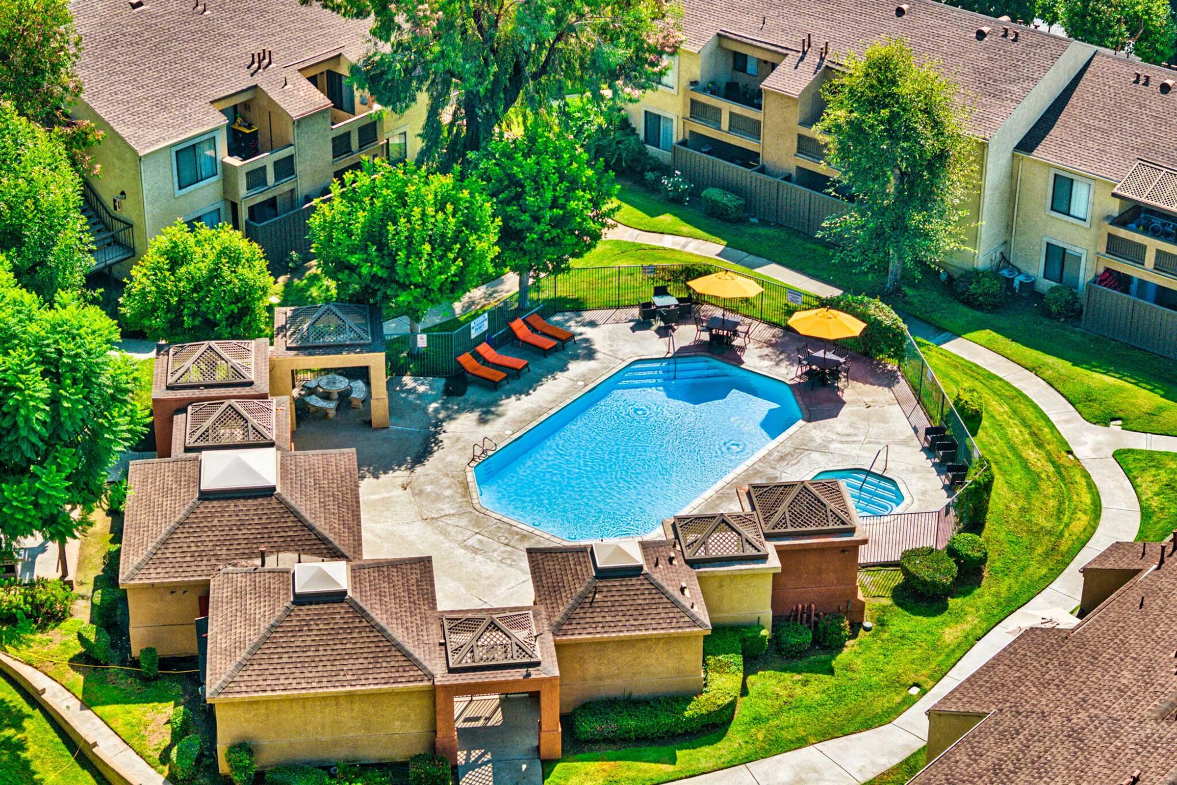 Aerial view of a residential complex featuring a swimming pool surrounded by lounge chairs and umbrellas. Lush green trees and landscaped pathways give a vibrant atmosphere. Multiple buildings in the background complete the scene, creating a serene outdoor living environment.