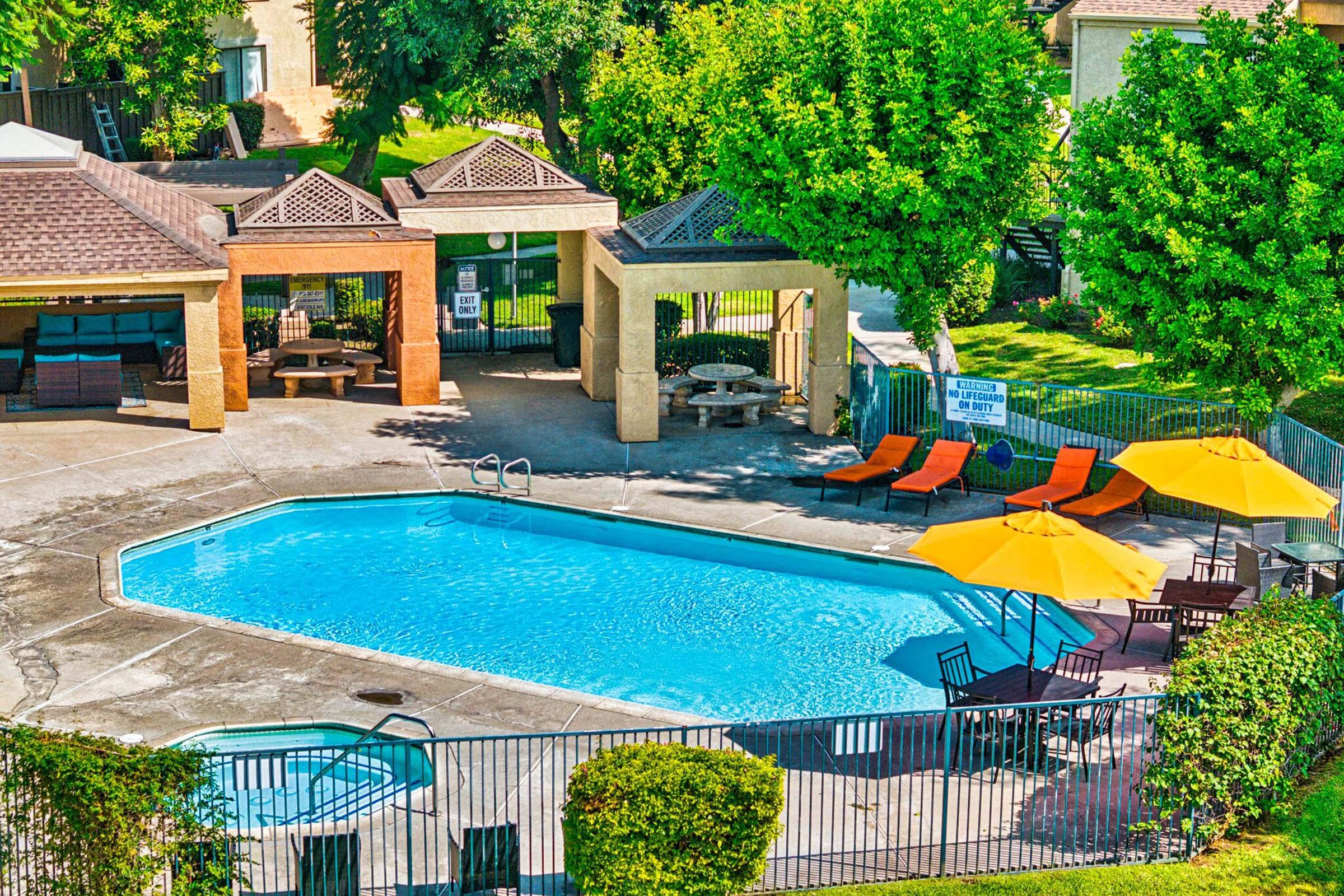 Aerial view of a residential pool area featuring a clear blue swimming pool, surrounded by orange lounge chairs and large yellow umbrellas. The area includes shaded seating under pavilions, with well-maintained grass and trees in the background. A sign can be seen near the poolside.
