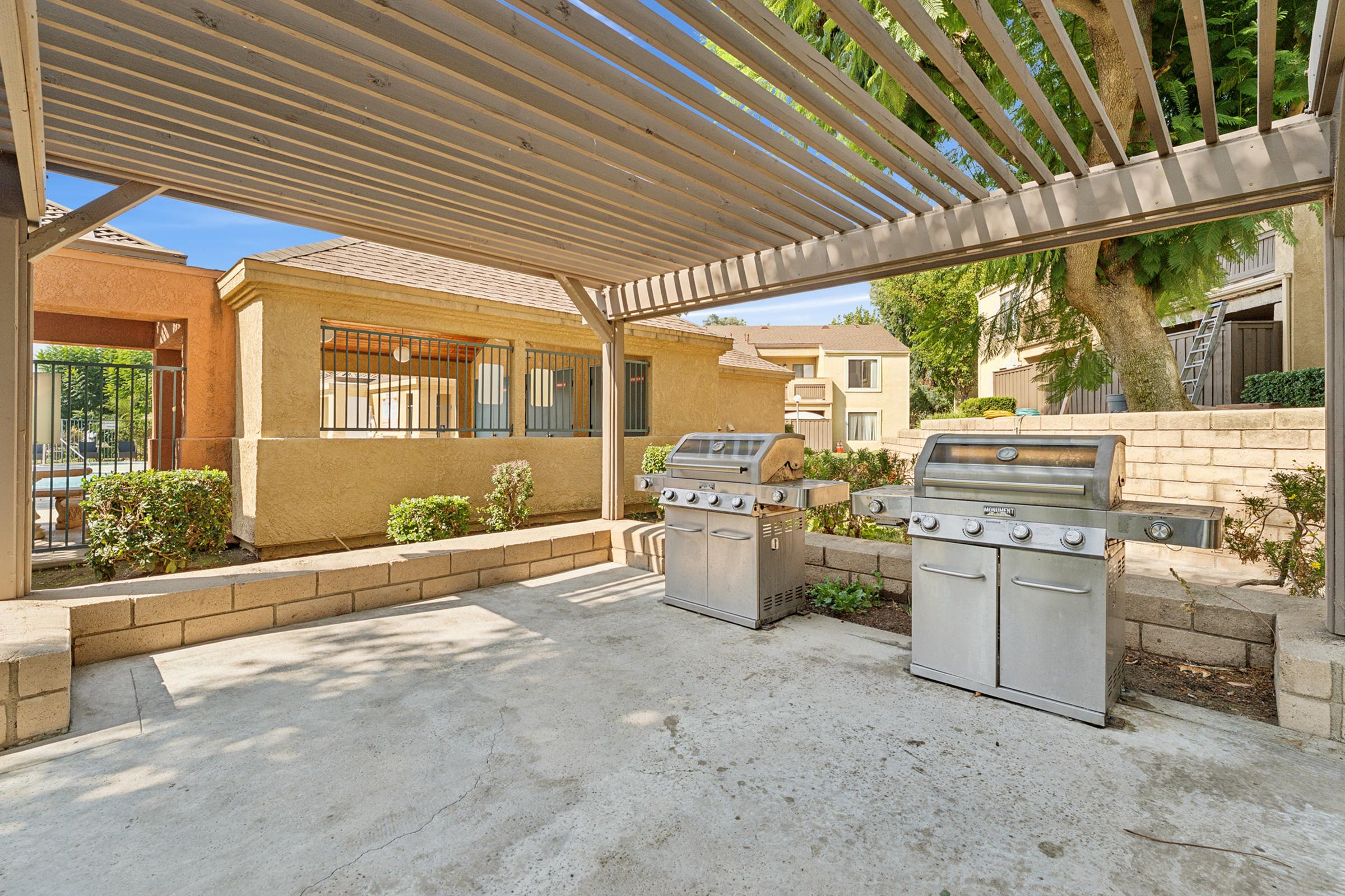 Outdoor grilling area featuring two stainless steel barbecue grills under a wooden pergola. Surrounding the area are neatly trimmed bushes and a landscaped yard. The setting is bright with clear blue skies, providing a welcoming space for outdoor cooking and gatherings.
