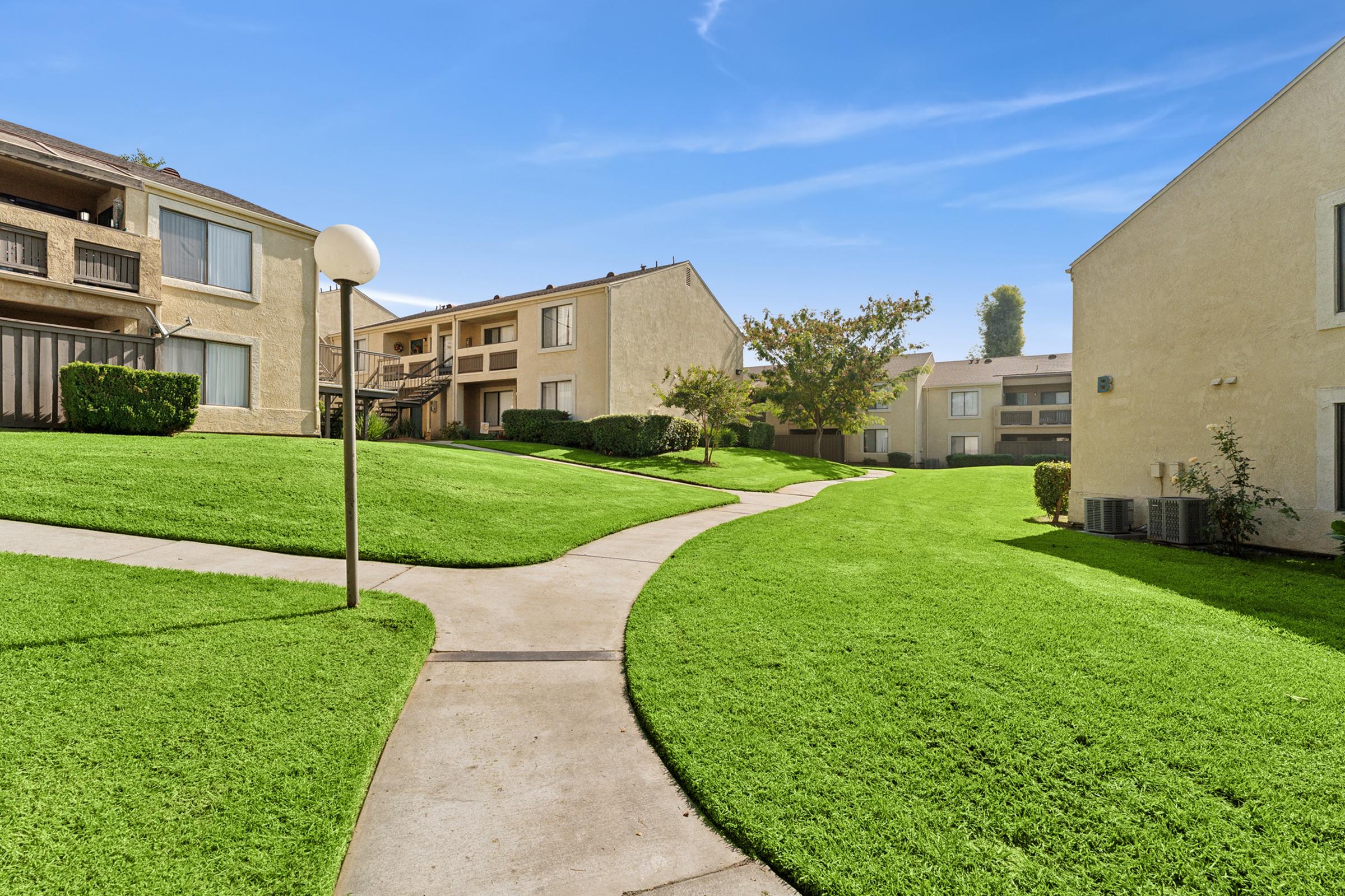 A landscaped area featuring apartment buildings, with well-maintained green grass and a winding pathway. The scene is set under a clear blue sky, creating a serene and inviting atmosphere. Decorative shrubs and trees enhance the appeal of the outdoor space.
