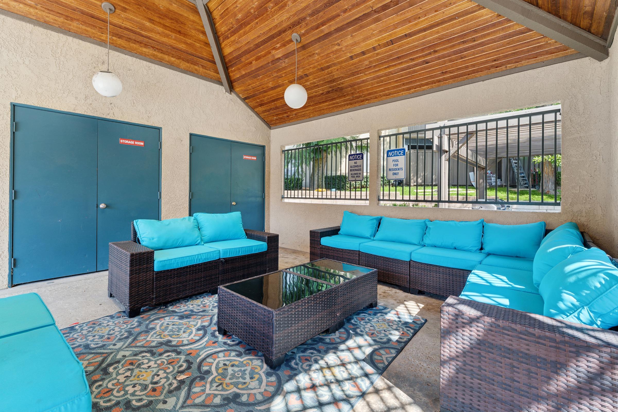 A cozy outdoor lounge area featuring turquoise cushions on dark wicker seating, a glass coffee table, and a colorful patterned rug. The space is sheltered by a wooden ceiling and includes blue doors on one side, with a view of greenery through a gated window.
