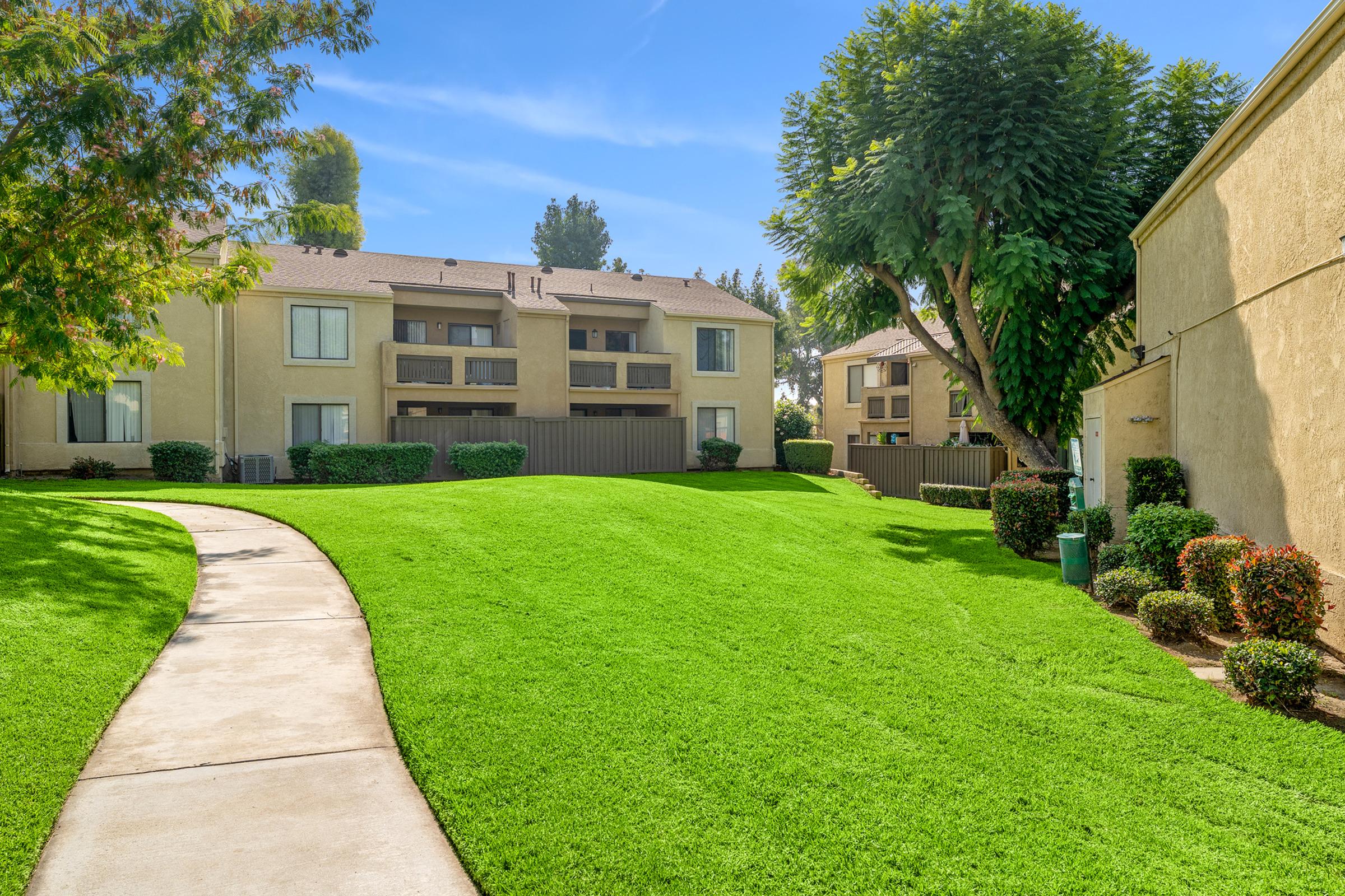 Lush green lawn with a concrete pathway leading through a well-maintained apartment complex. The buildings feature balconies, surrounded by neatly trimmed shrubs and trees under a clear blue sky.