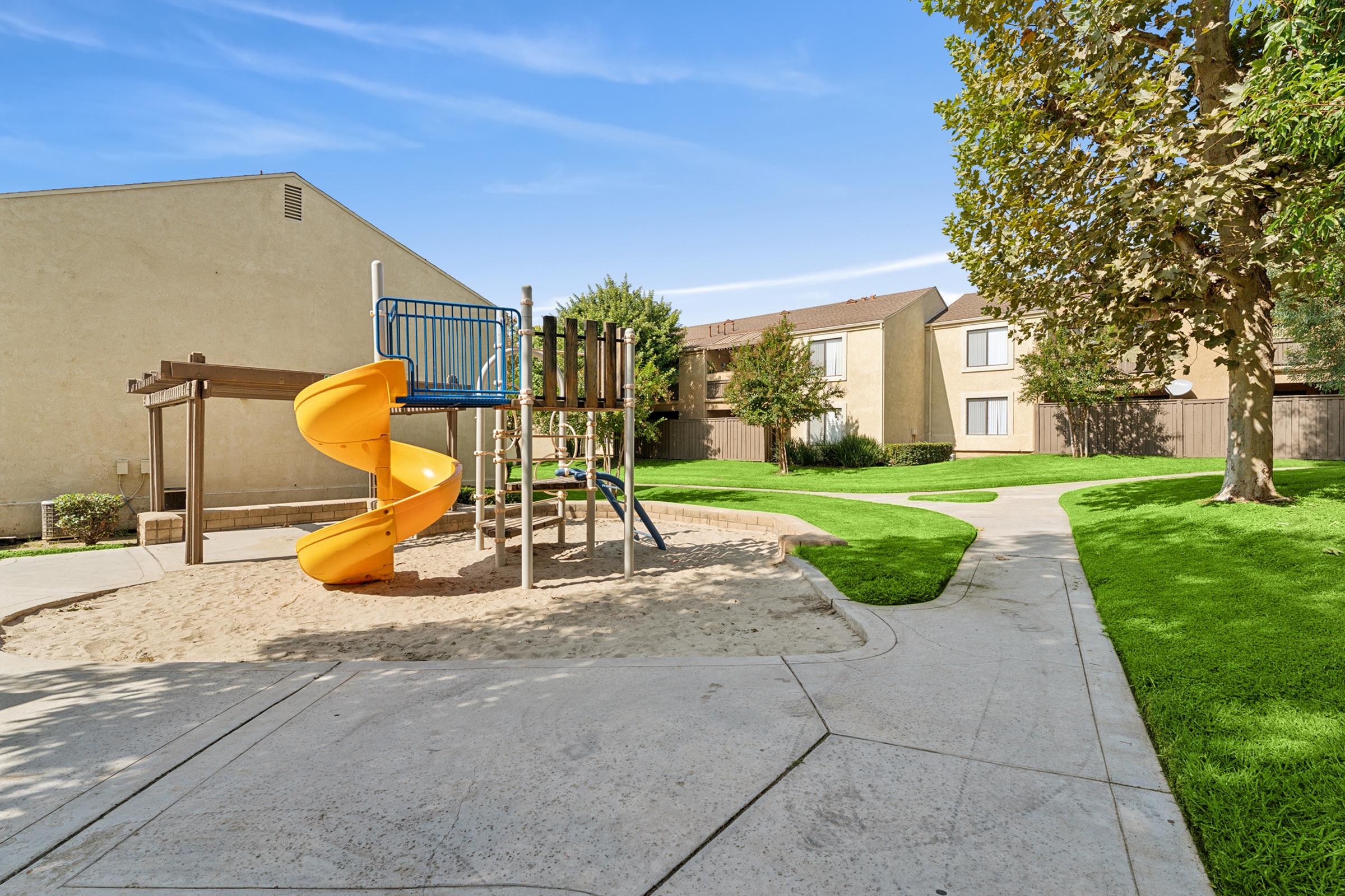 Playground area with a yellow slide, climbing structure, and sandbox. Surrounding grassy areas and trees. Apartment buildings are visible in the background under a clear blue sky.