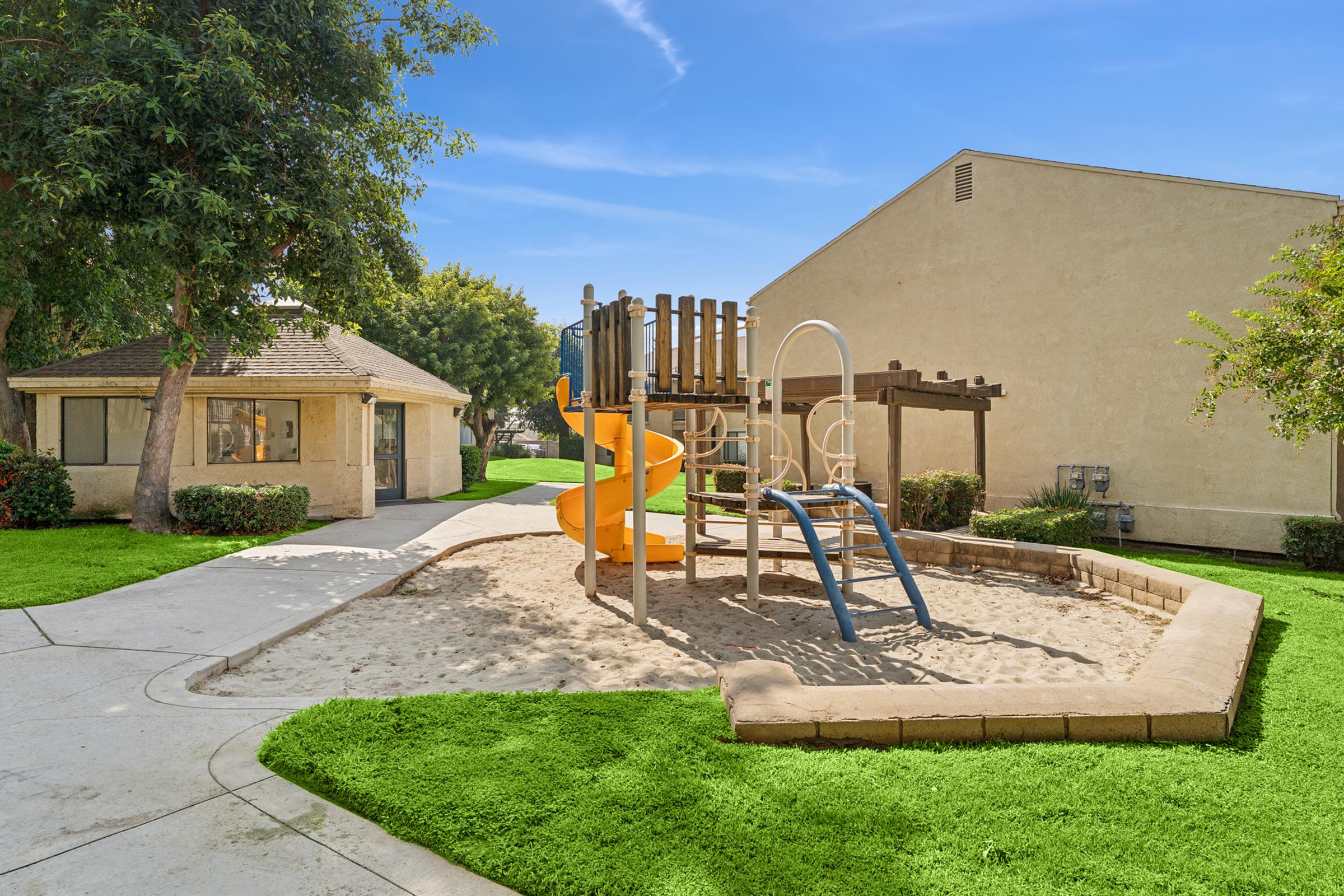 A playground featuring a yellow slide and play structure set in a sandy area, surrounded by green grass. Nearby, there is a walkway connecting to a building with a beige exterior and a sloped roof. Trees provide shade in the background, creating a sunny, inviting outdoor space for children.