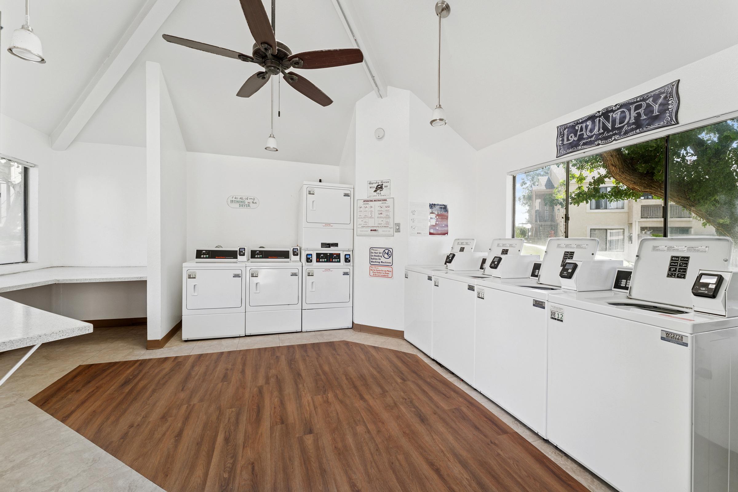 A clean and spacious laundry room with several white washing machines and dryers lined up against the wall. The room features a high ceiling with a ceiling fan, large windows letting in natural light, and a laundry sign on the wall. The floor is covered with light brown laminate.