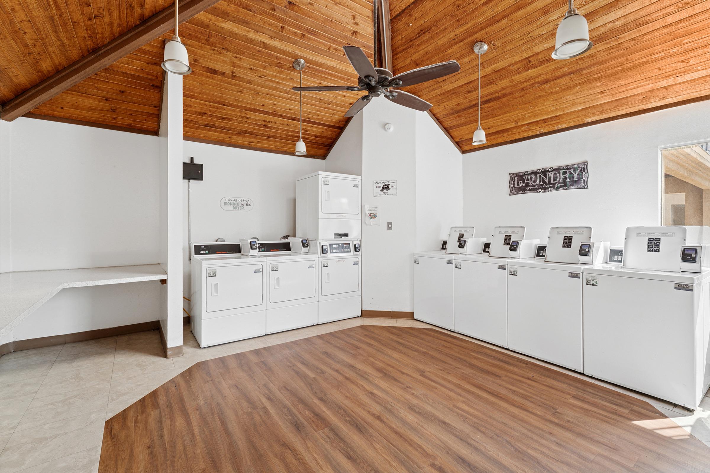 A modern laundry room featuring several white washing machines and dryers arranged along one wall. The space has a wooden ceiling with exposed beams, a ceiling fan, and a light-colored floor. A folding table and wall signage indicate the laundry area. Natural light may filter in from windows.