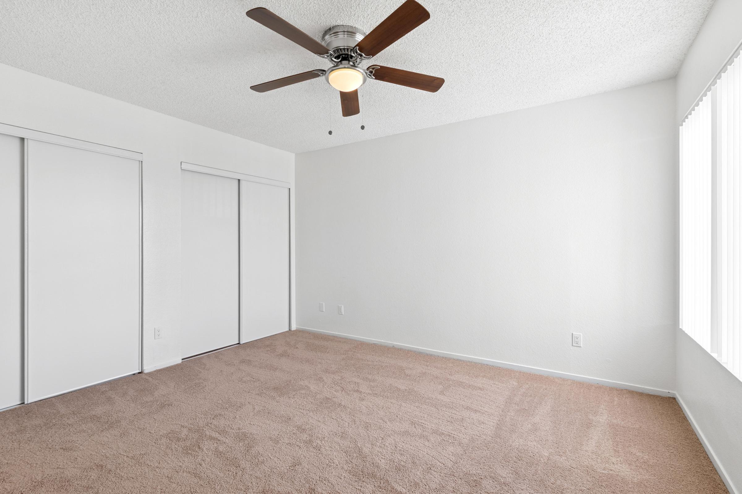 A spacious, empty room with beige carpet and white walls. It features a ceiling fan with wooden blades, two closed white closet doors, and a large window allowing natural light to fill the space. The room has a clean, minimalistic look and is ready for furnishings.
