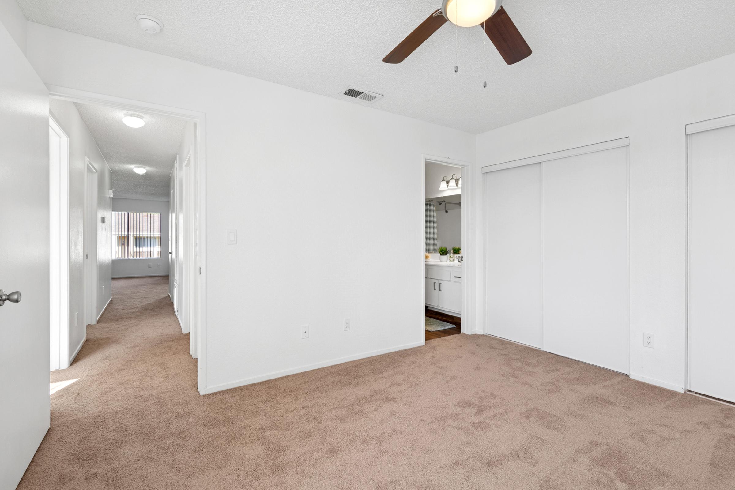 Interior view of a light-colored room featuring beige carpet, white walls, and a ceiling fan. Two doorways lead to adjacent spaces, with one doorway opening into a bathroom area visible in the background. The room has ample natural light, creating a bright and inviting atmosphere.