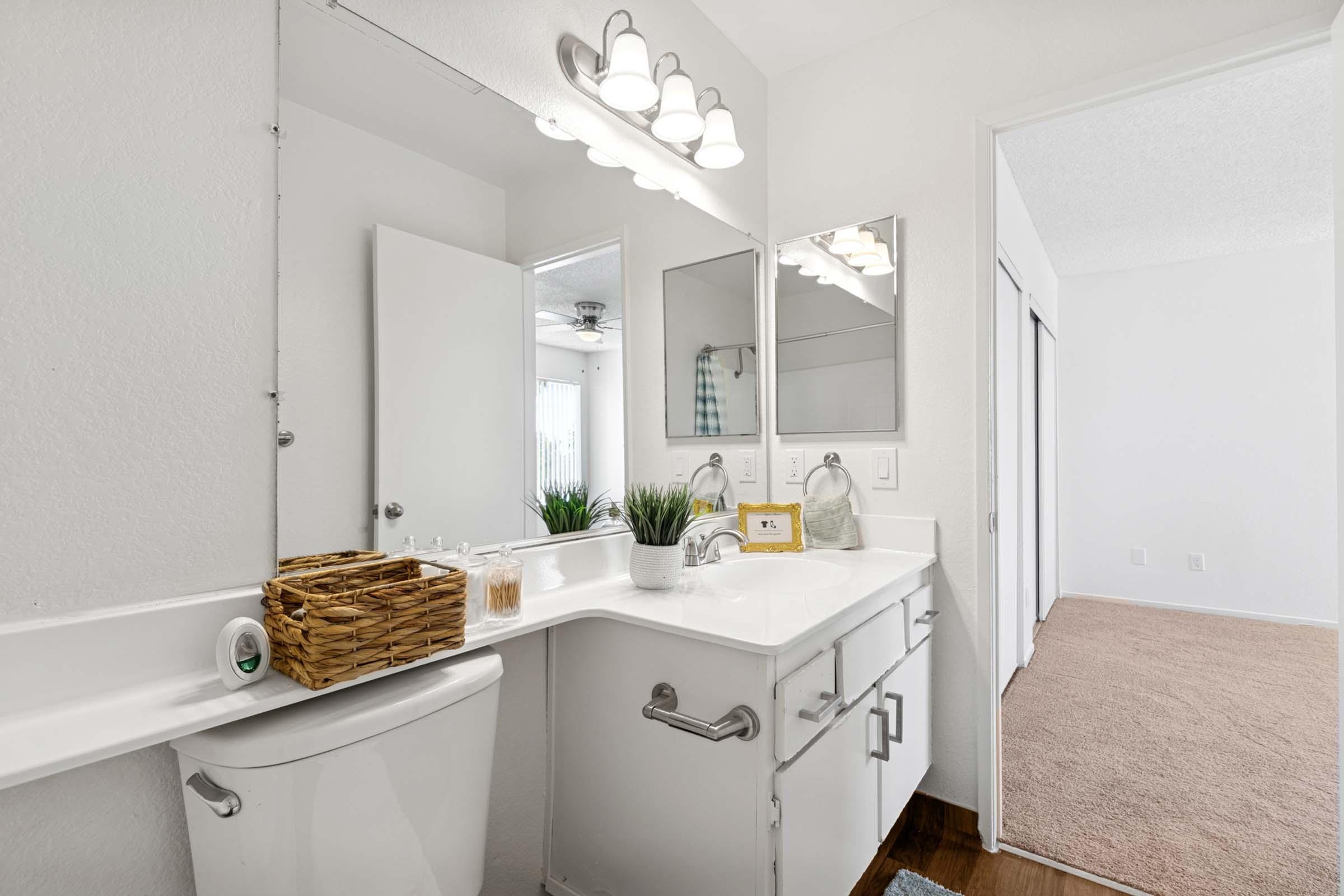 A clean and modern bathroom featuring a white vanity with two mirrors and a double sink. There’s a wall-mounted light fixture above the mirrors, a decorative basket on the counter, and a small plant. A door leads to another room, and soft, neutral-colored carpeting is visible on the floor.