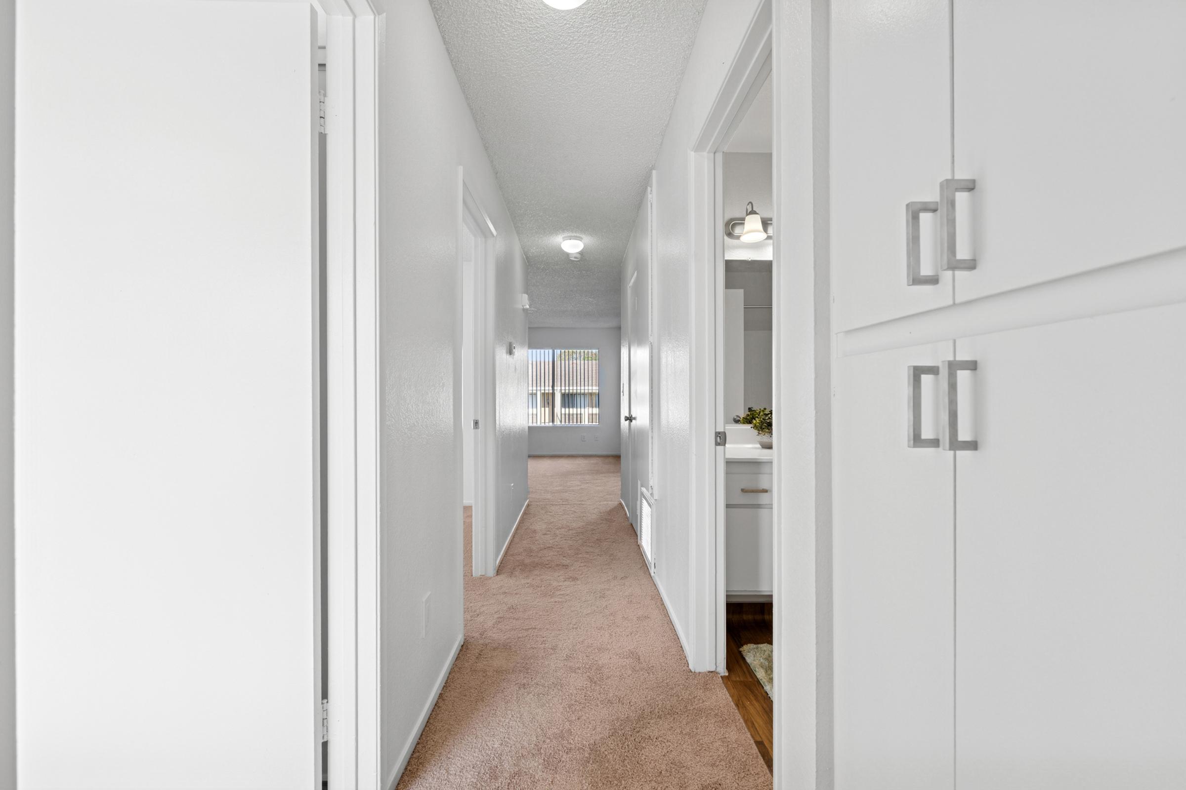 A well-lit hallway with beige carpet and white walls. The view includes closed doors on either side, leading to rooms, with a glimpse of a light-filled space at the end. The doors feature simple handles, and there are light fixtures on the ceiling providing illumination.
