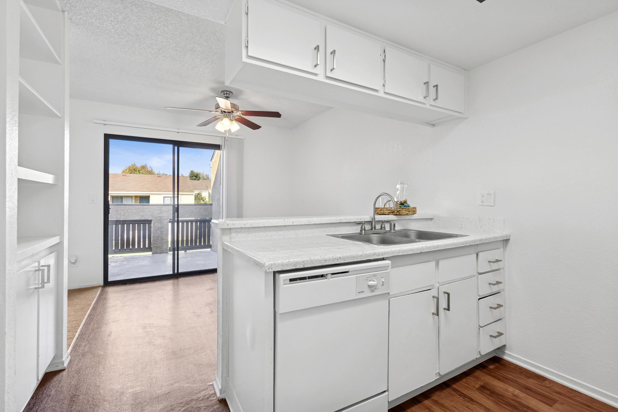 A modern kitchen with white cabinetry, a double sink, and a dishwasher. The countertop is light-colored, and there's a ceiling fan above. A sliding glass door leads to a balcony, allowing natural light to enter the room. The flooring is a warm wood tone, creating a cozy atmosphere.