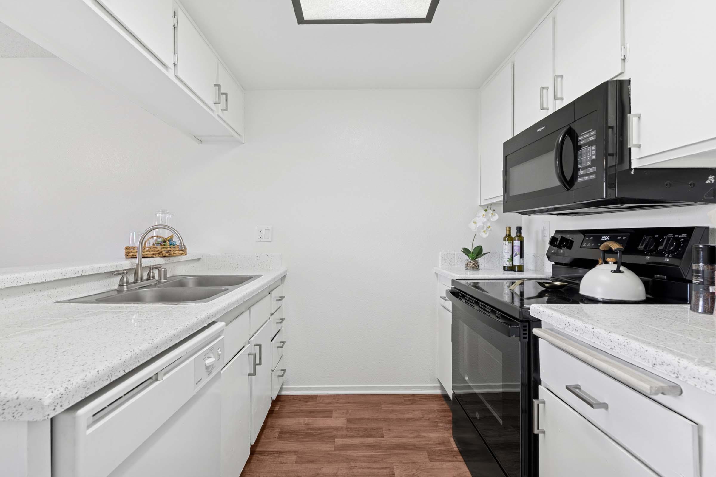 A modern kitchen featuring white cabinetry, a double sink with a polished countertop, a black microwave above the stove, and a sleek black oven. The flooring is a warm wood tone, and there are decorative items like a plant and a bottle of oil on the counter. Natural light illuminates the space.
