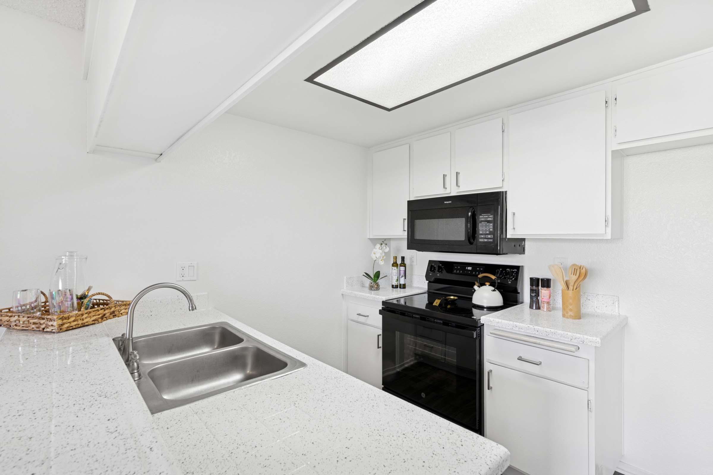 A modern kitchen featuring white cabinets, a black stove and microwave, and a sleek white countertop with a double sink. The space is bright, with overhead lighting and a decorative basket on the counter, along with cooking utensils and a small plant.