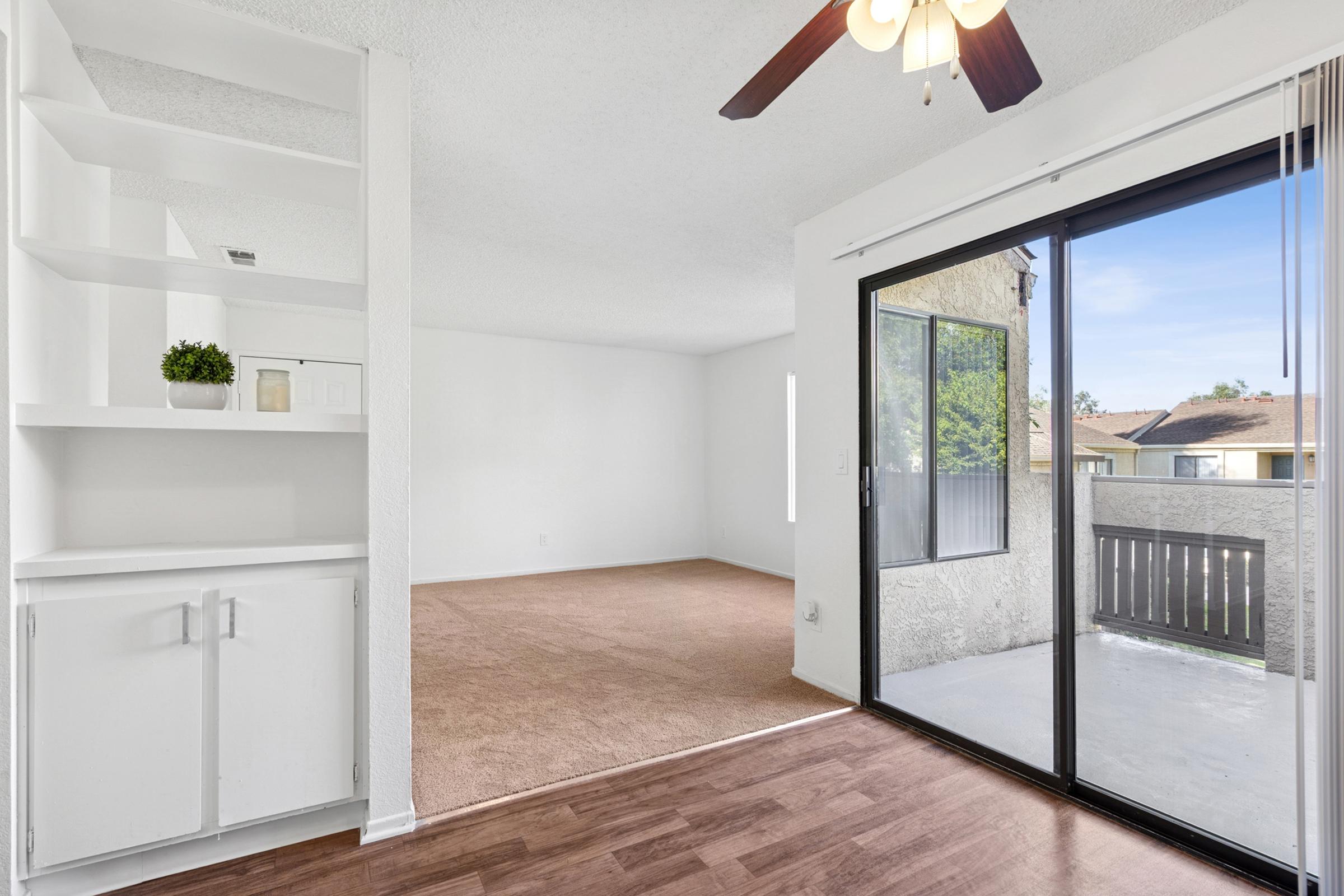 A bright, empty living space with light-colored walls and carpet. There is a ceiling fan, a built-in white bookshelf, and a large sliding glass door leading to a balcony with views of greenery outside. Natural light enters the room, creating an airy atmosphere.