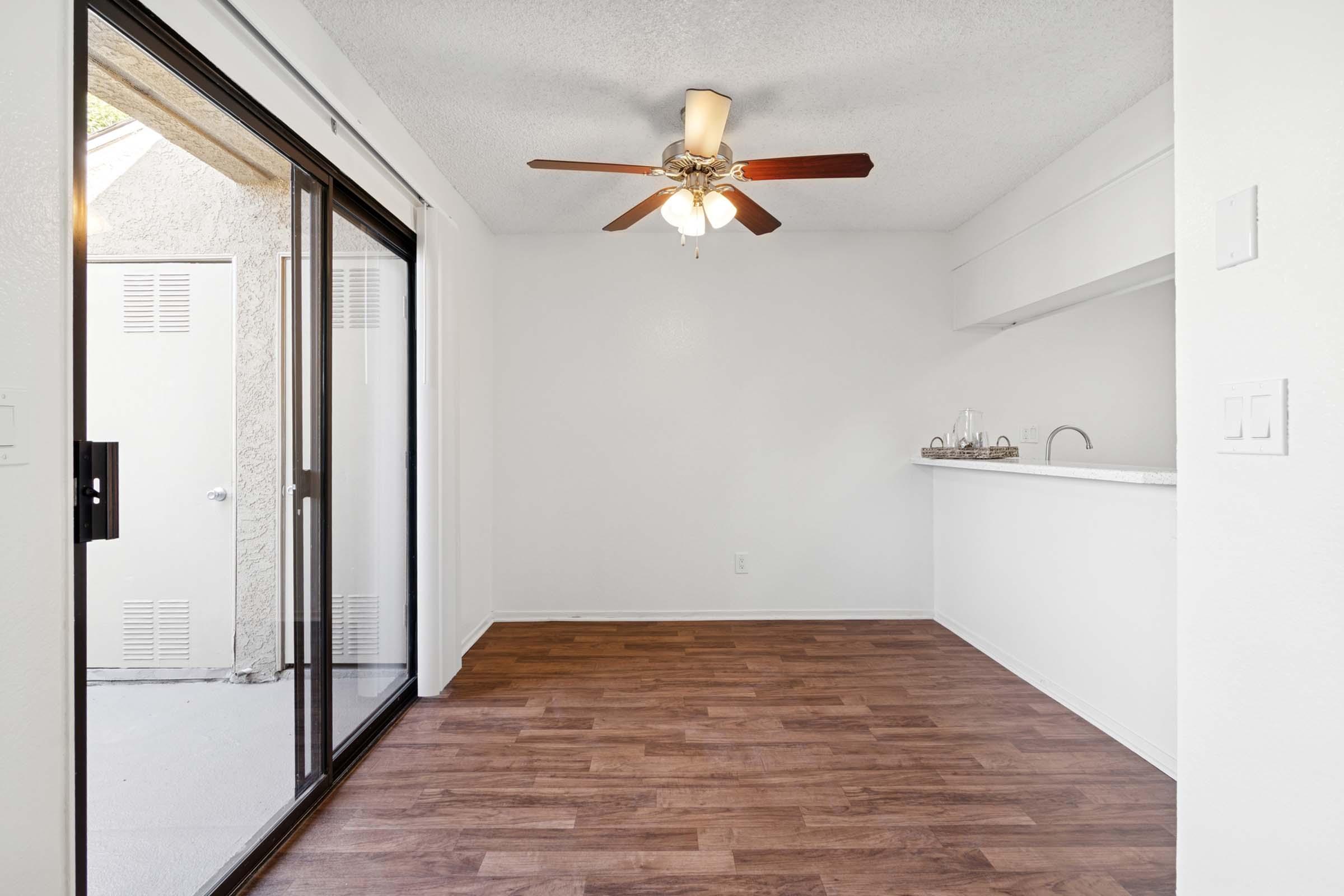 A brightly lit, empty room featuring a ceiling fan, hardwood-style flooring, and a sliding glass door leading to an outdoor area. The walls are painted white, and there is a small kitchen countertop visible in the background.