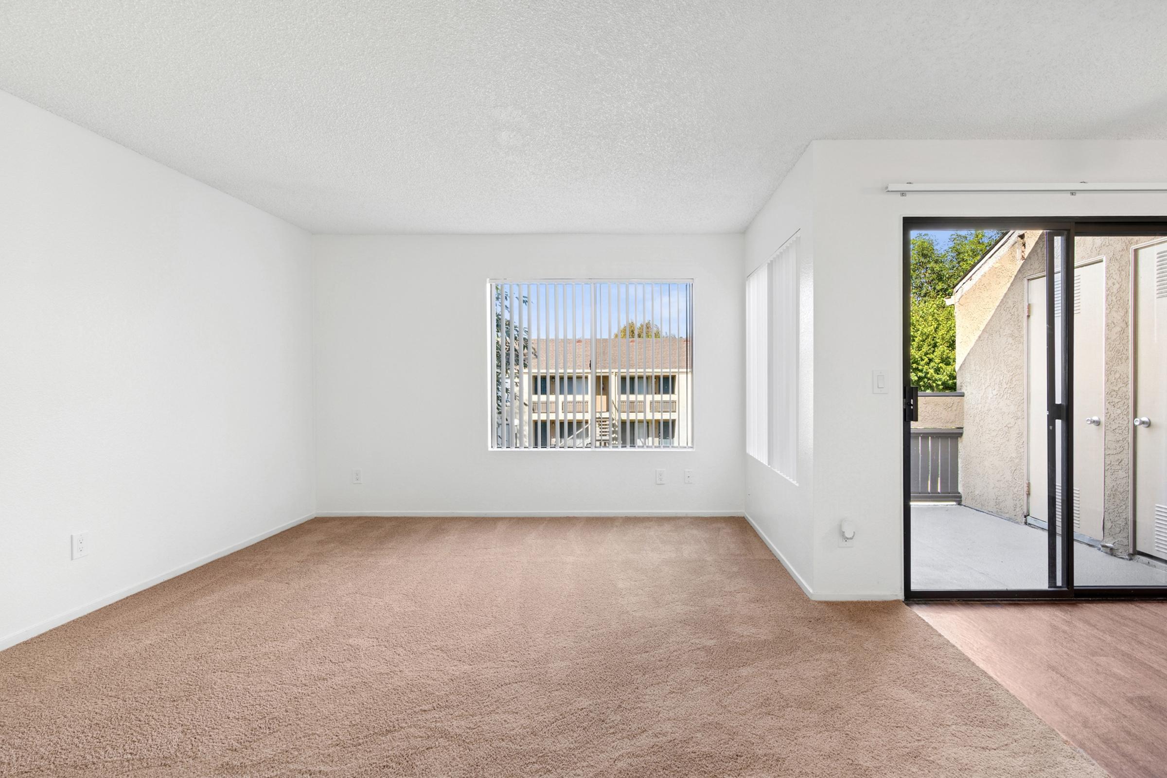Spacious interior of a room featuring beige carpeting, white walls, and a large window with vertical blinds. Natural light illuminates the space, showcasing an open area leading to a sliding glass door that opens to a balcony, creating a bright and inviting atmosphere.
