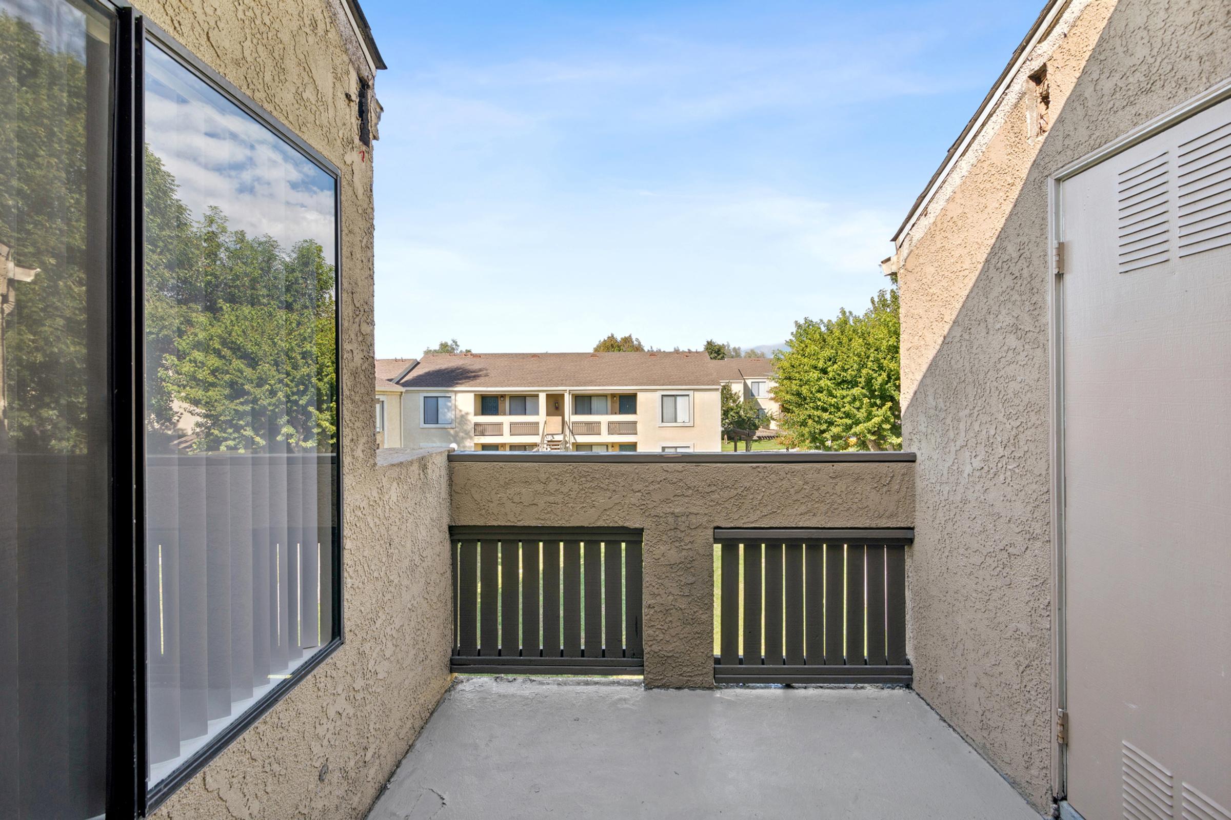 View from a small balcony or patio showing a residential area with green trees and a building across the way. The walls are textured and a large window is visible on one side, providing natural light.