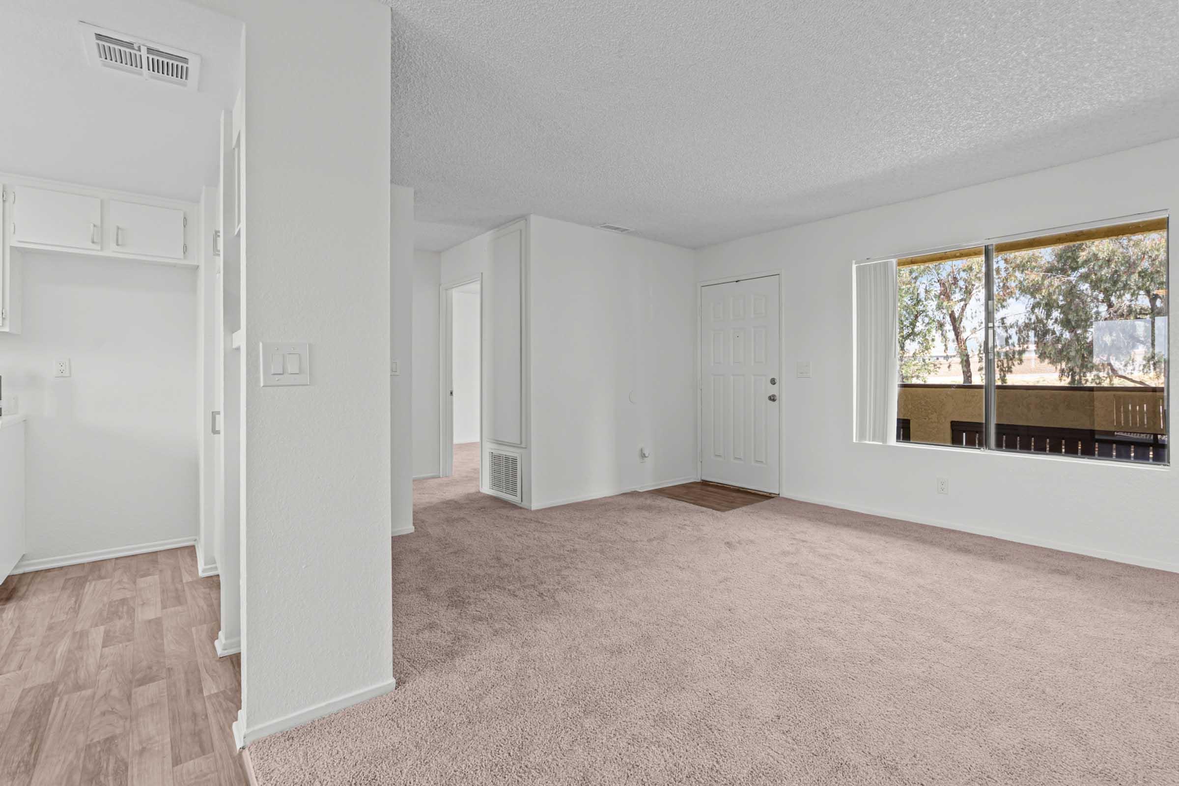 Spacious, empty living area featuring light-colored walls and carpet. A large window lets in natural light, revealing views of trees outside. The layout includes a doorway leading to another room and a kitchen area visible to the left.