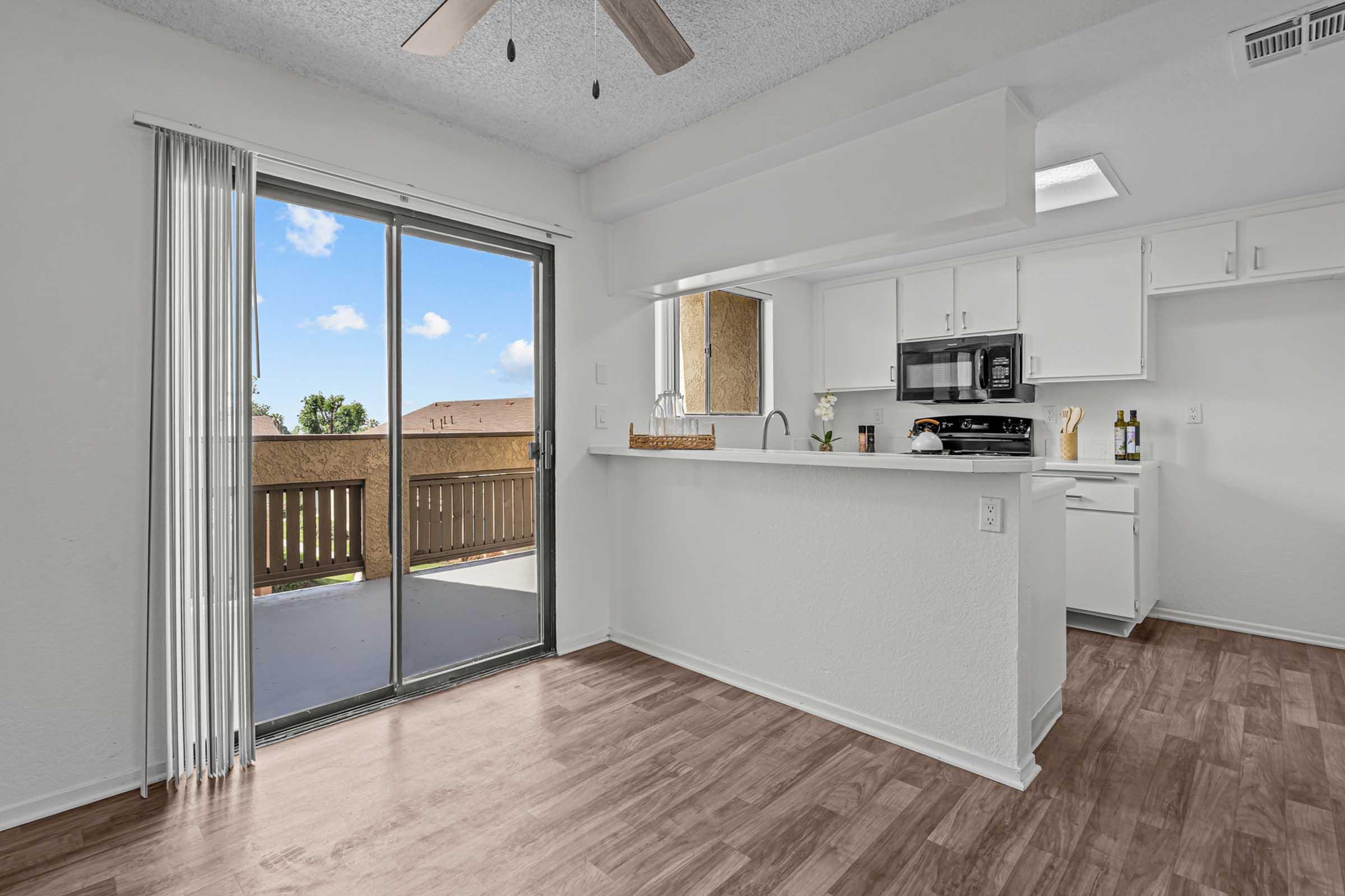 Bright kitchen and dining area with white cabinetry and modern appliances. A sliding glass door leads to a balcony with outdoor access. The flooring is a light wood finish, and the space is filled with natural light, creating an inviting atmosphere.