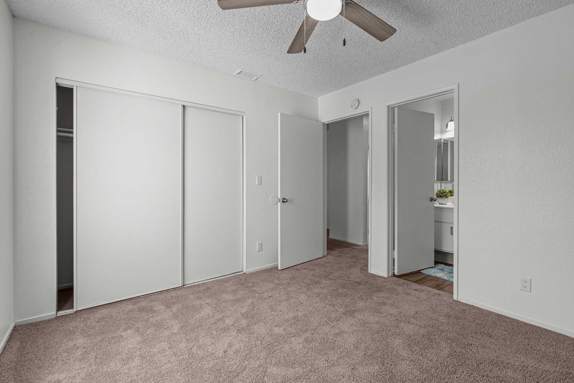 Empty room with beige carpet and white walls. It features a ceiling fan, a closet with sliding doors, and two doorways leading to other rooms. The natural light creates a bright and open atmosphere, providing a spacious feel to the area.