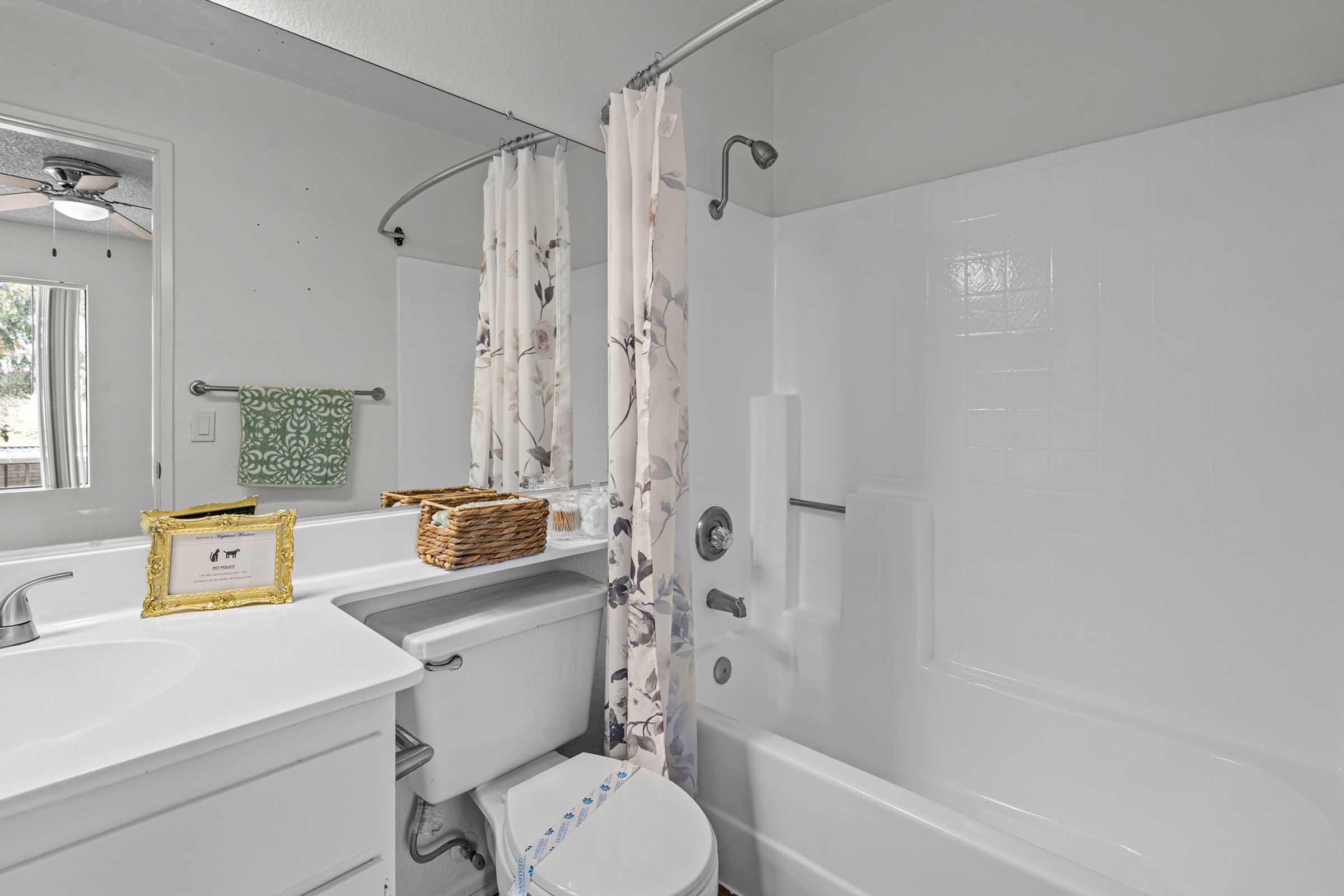 A clean, modern bathroom featuring a white bathtub, shower with glass doors, a floral shower curtain, a white vanity with storage, and a decorative basket. Natural light comes in from a window, enhancing the bright and airy atmosphere.