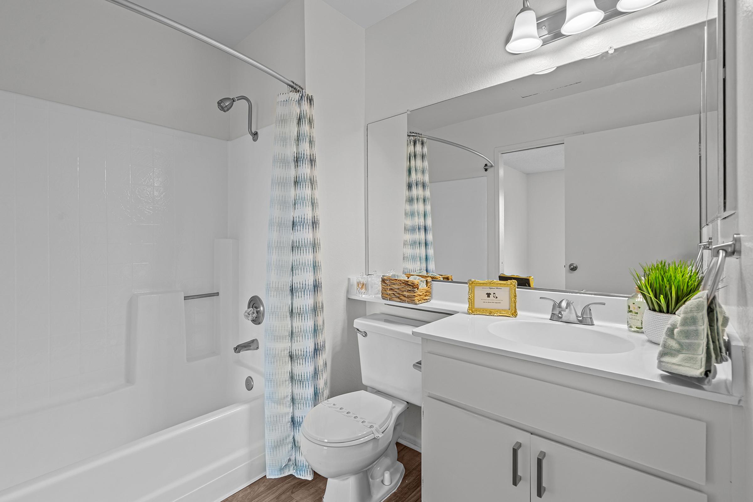 A clean and modern bathroom featuring a white bathtub with a shower, a large mirror above the sink, and a countertop with decorative items. The walls are painted a light color, and there is a window with blue and white checkered curtains. The floor has a wood-like appearance.