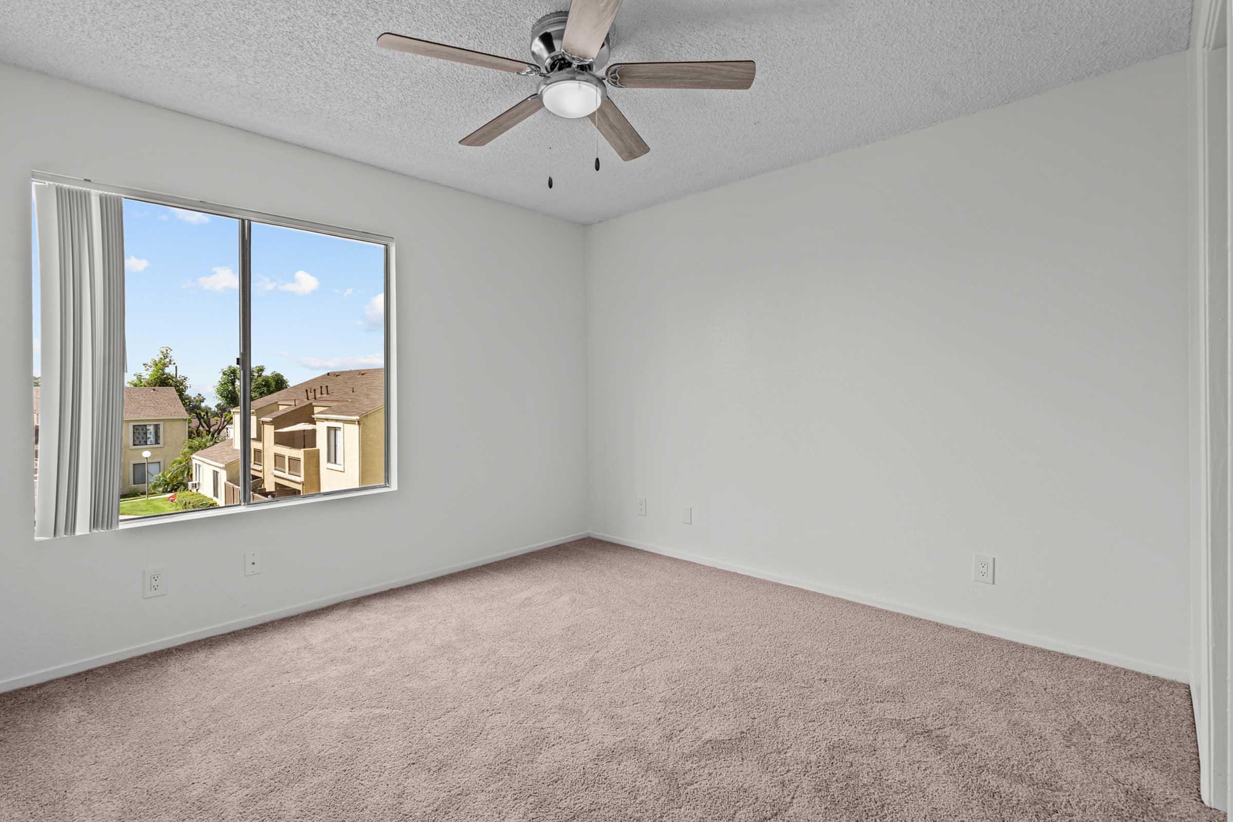 A spacious, empty room with light-colored walls and a ceiling fan. It features a large window with a view of nearby buildings and greenery, providing natural light. The floor is covered in plush beige carpeting, enhancing the bright and airy feel of the space.